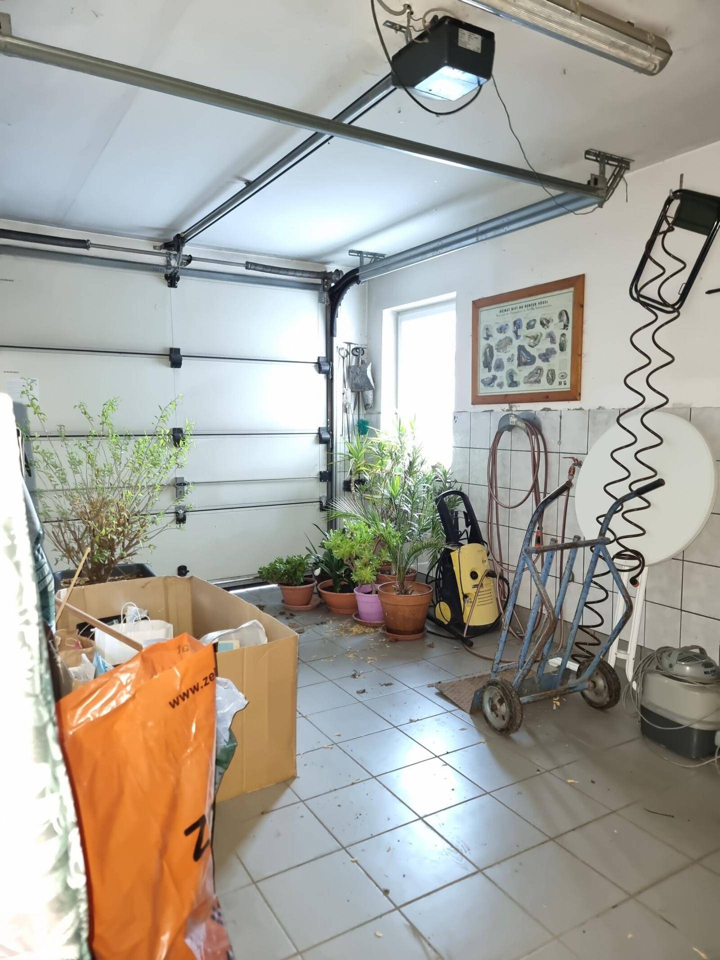 Garage mit Fliesenboden, Fenster und Abstellmöglichkeiten für Gartengeräte und Pflanzen.