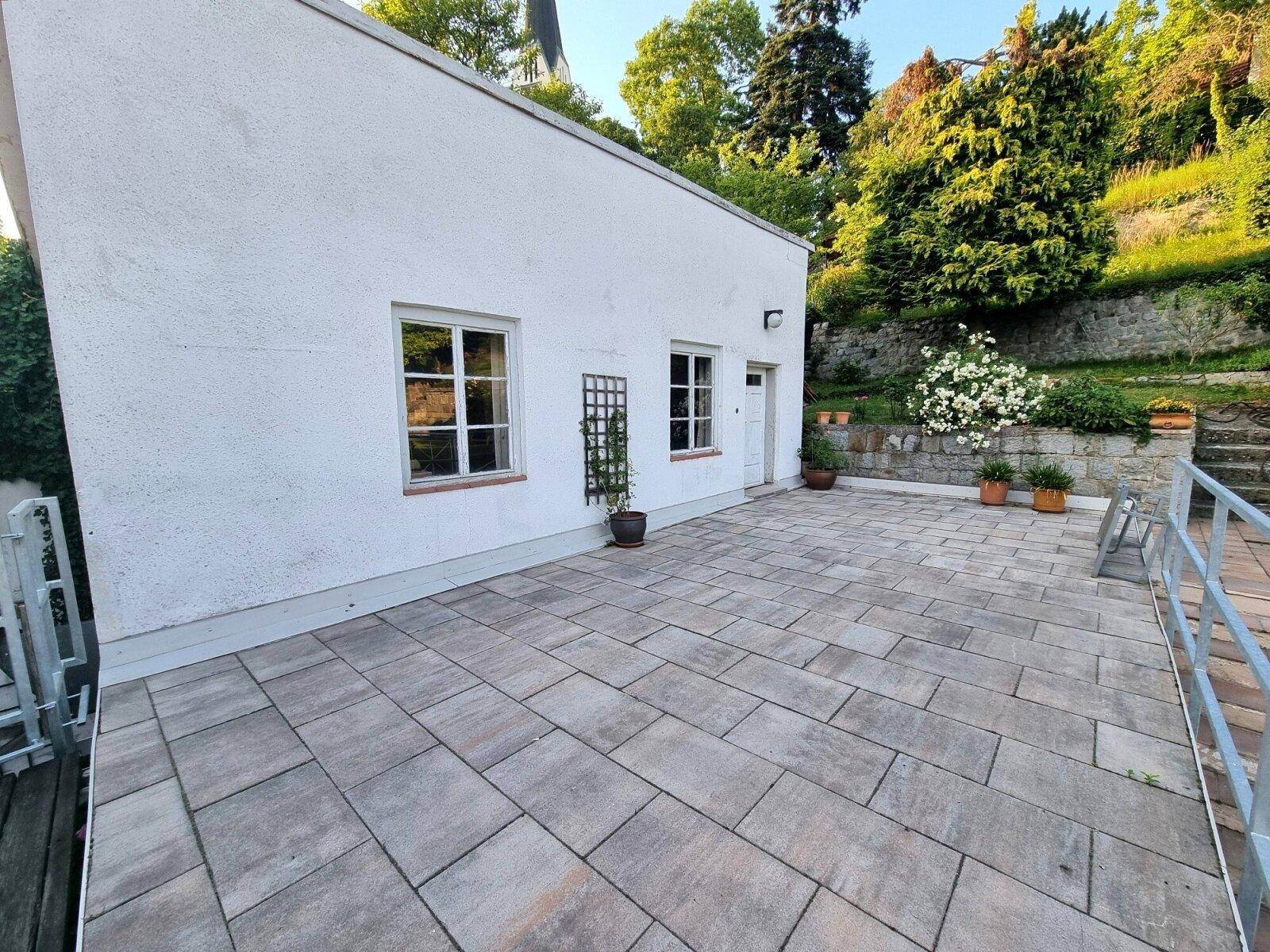 Große, gepflasterte Terrasse neben einem weißen Gebäude mit Blick auf einen bewachsenen Hang.