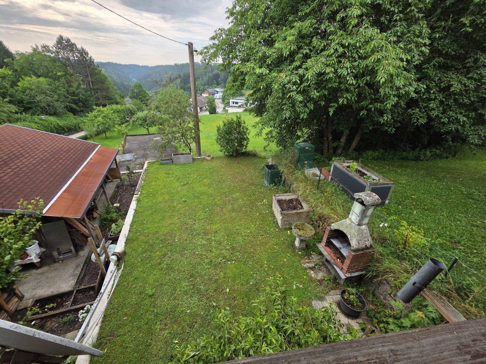 Gartenbereich mit Rasenfläche, gemauertem Grill und Hochbeeten, umgeben von grüner Natur.