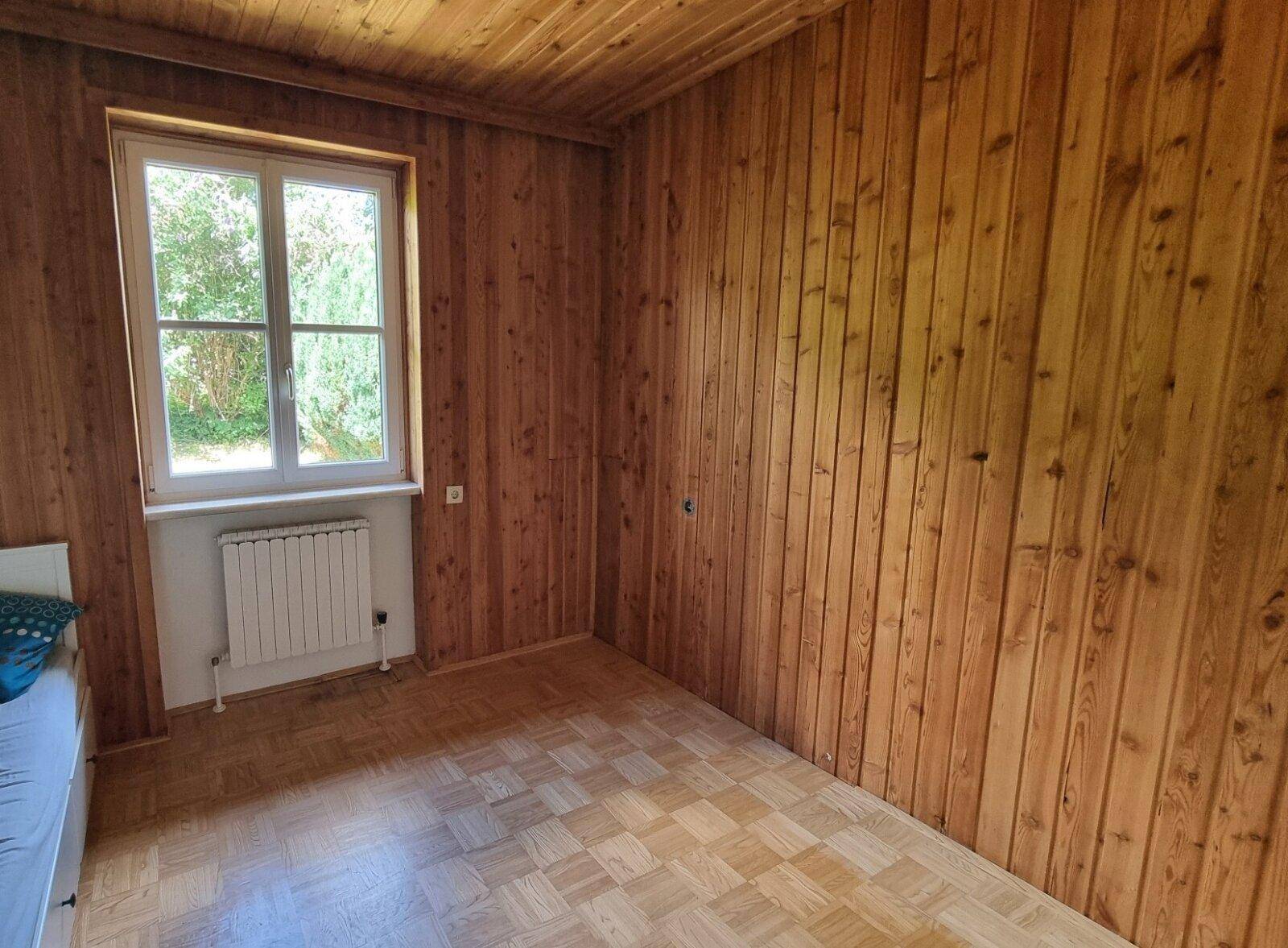 Schlafzimmer mit Holzwänden, Holzdecke und Parkettboden, hell durch ein Fenster.