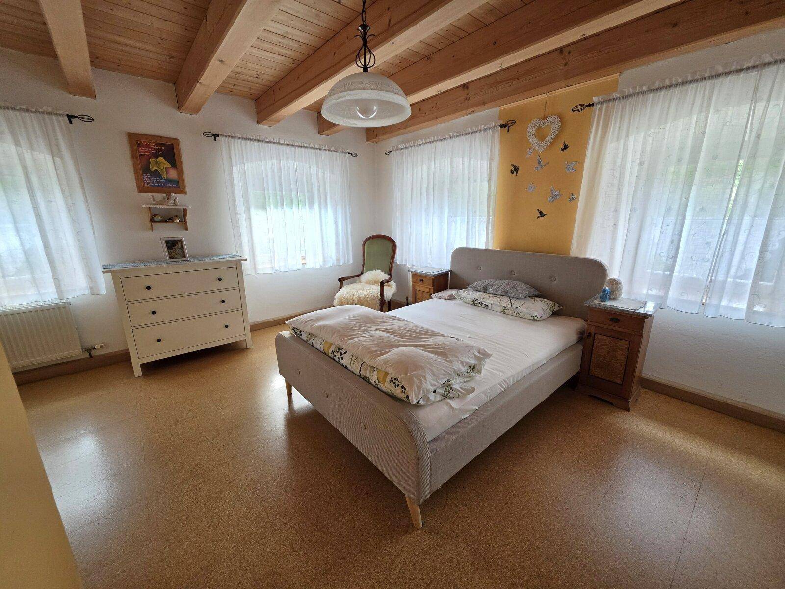 Schlafzimmer mit Holzbalkendecke, gemütlichem Bett und viel Tageslicht durch zwei Fenster.