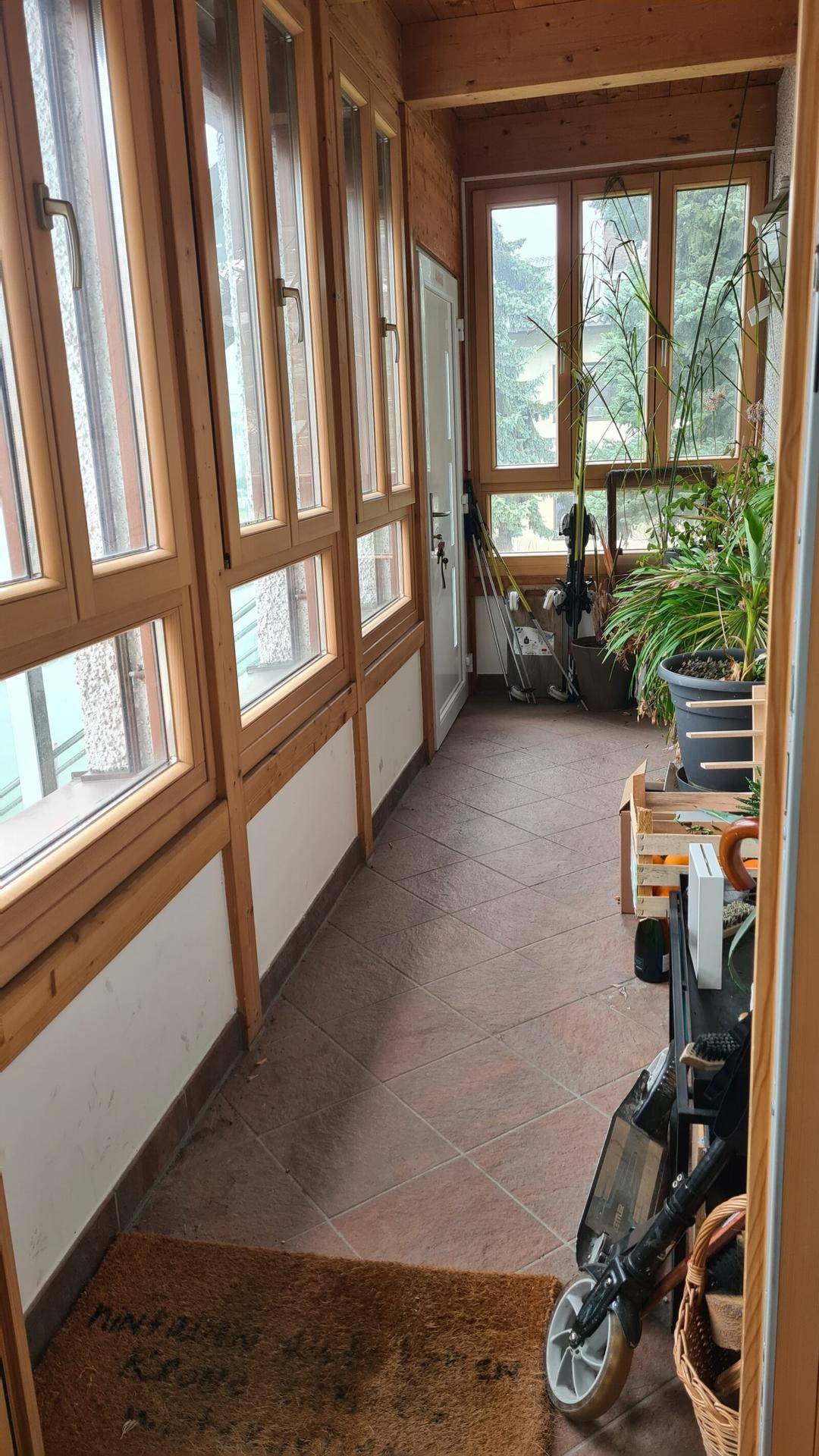 Heller Wintergarten mit vielen Holzfenstern, Fliesenboden und Pflanzen.