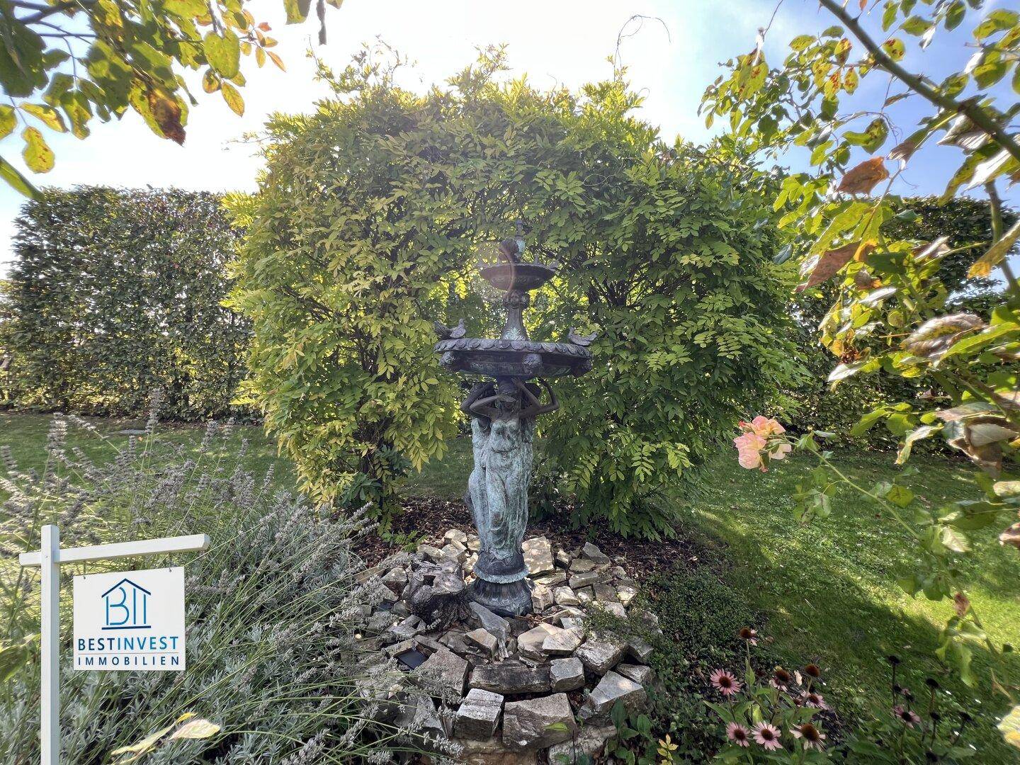 Dekorativer Brunnen im Garten, umgeben von üppigem Grün und blühenden Sträuchern.