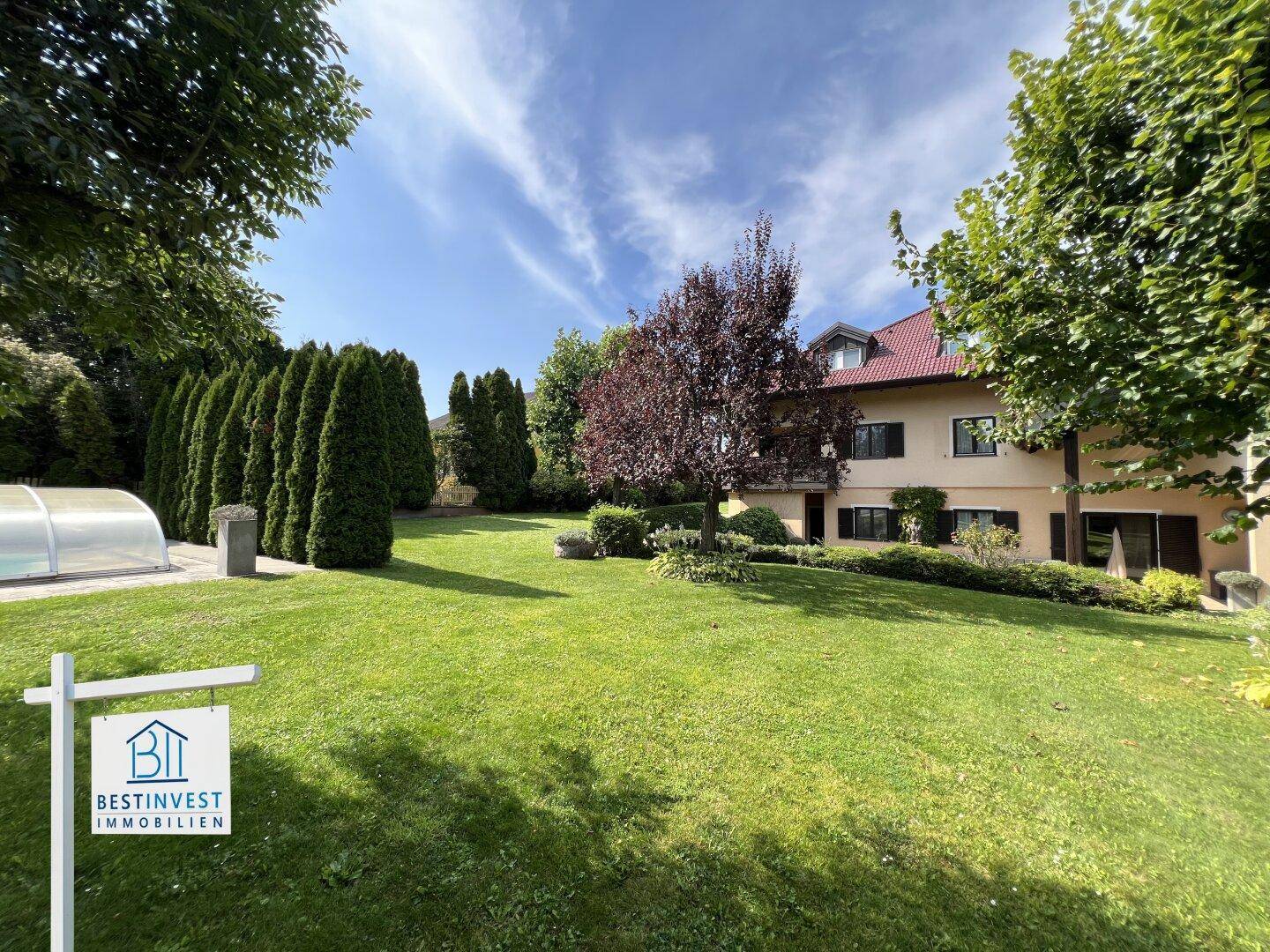 Weitläufiger Garten mit Rasen, Bäumen und dem Wohnhaus im Hintergrund unter blauem Himmel.