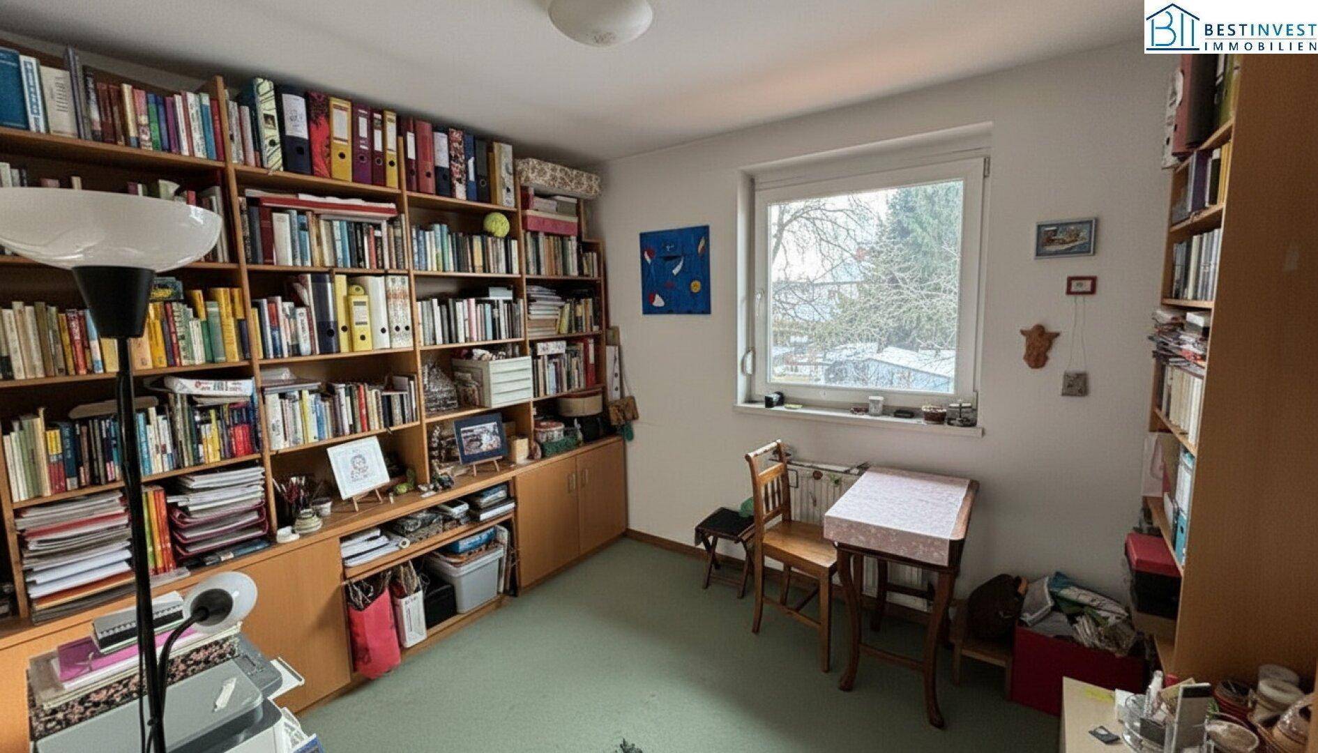 Arbeitszimmer mit großen Bücherregalen, Teppichboden und Fenster mit Blick ins Grüne.