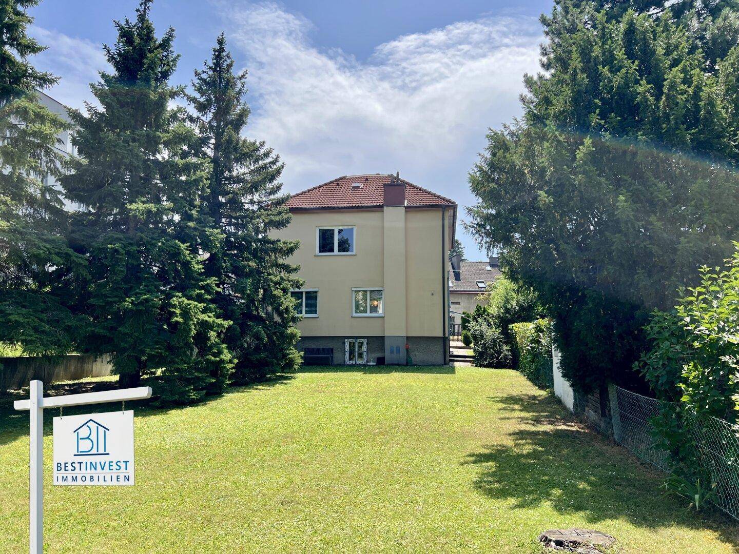Einfamilienhaus mit großem Garten und viel Grünfläche umgeben von Bäumen unter blauem Himmel.