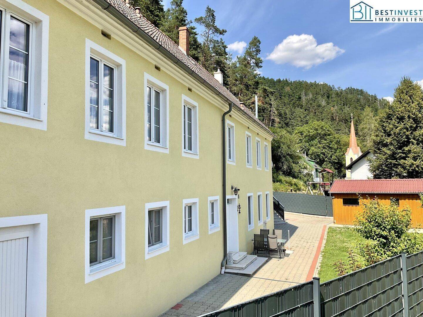 Seitenansicht des gelben Gebäudes mit gepflastertem Weg, Rasenfläche und Blick auf eine Kirche im Hintergrund.