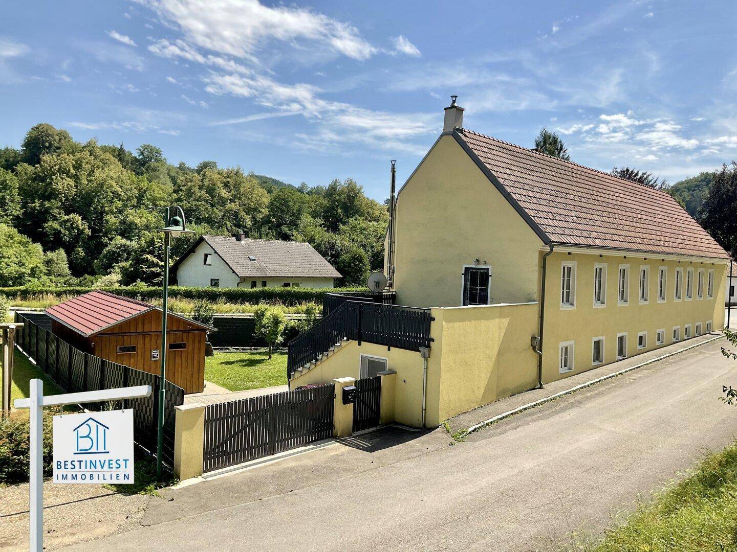 Seitenansicht des gelben Gebäudes mit Satteldach, umgeben von grünen Hügeln und einem kleinen Schuppen.