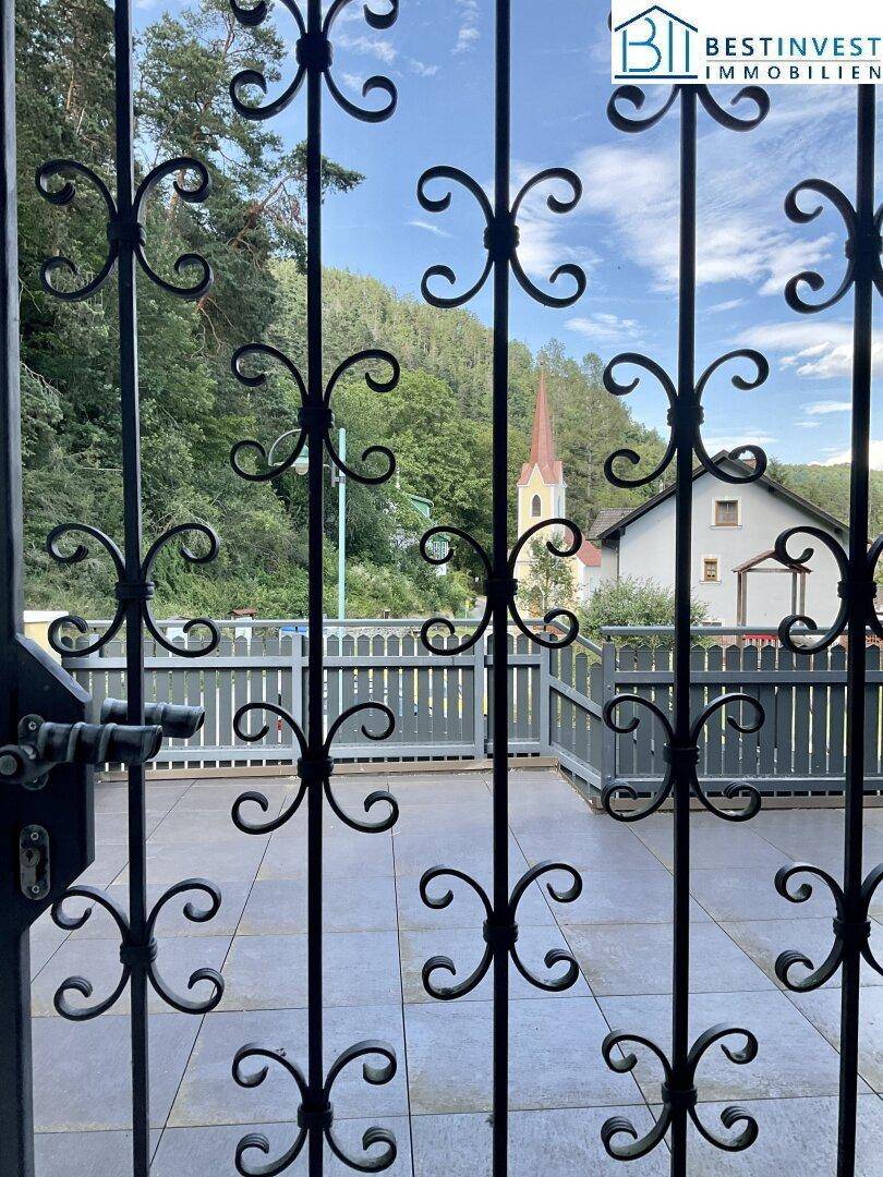 Blick durch ein verziertes Metalltor auf eine Terrasse mit Holzzaun und eine Kirche im Hintergrund.