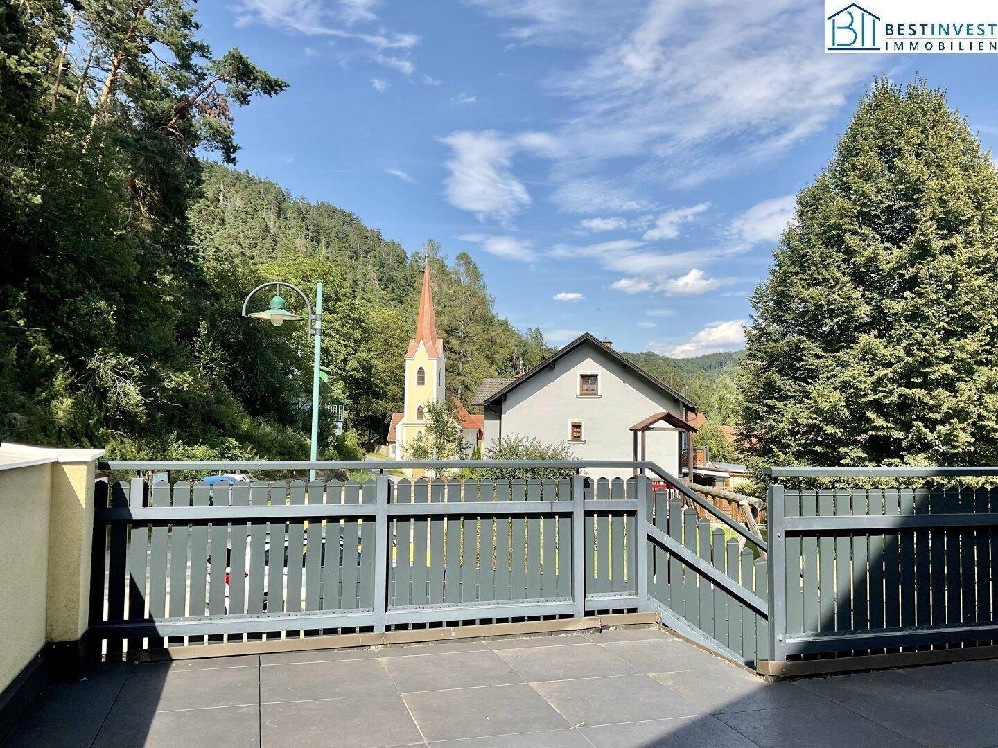 Geräumige Terrasse mit Holzzaun und weitem Blick auf die umliegende Landschaft und eine Kirche.