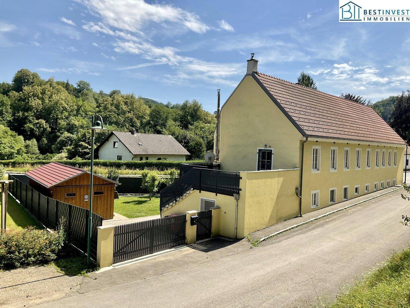 Gesamtansicht des gelben Gebäudes mit Satteldach, umgeben von grüner Landschaft und einem Nebengebäude.