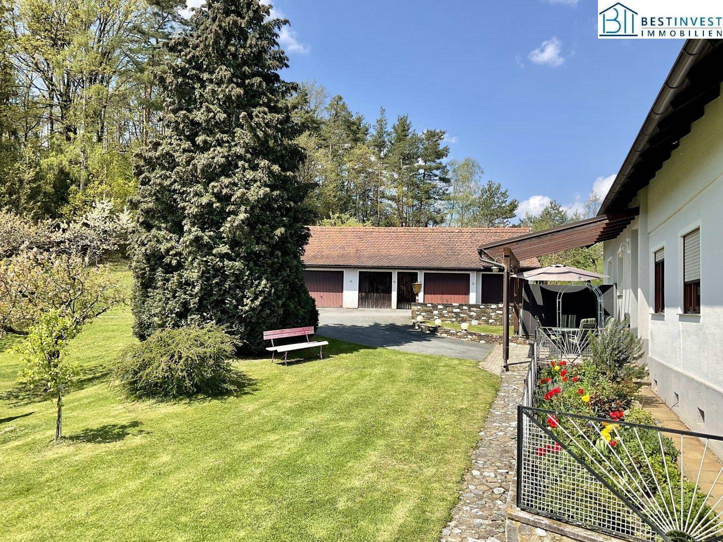 Gepflegter Gartenbereich mit Rasenfläche, hohen Bäumen und einer Sitzbank, im Hintergrund eine Garage.
