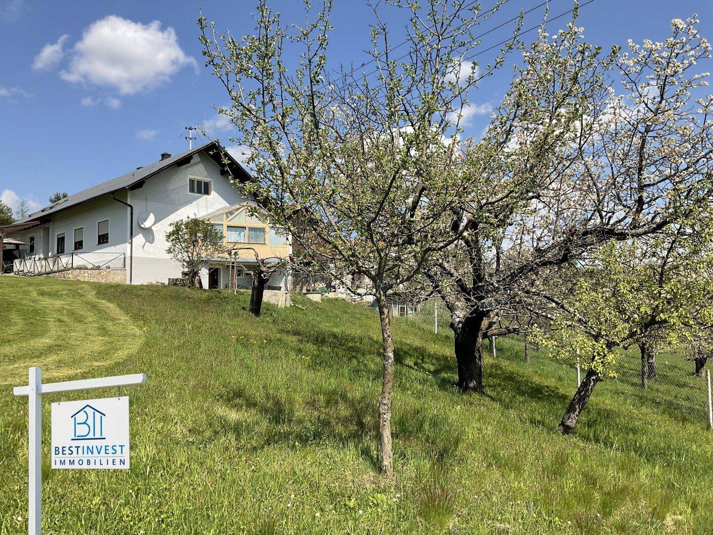Haus auf einem Hügel mit Obstbäumen und einem Wintergarten, umgeben von Natur.