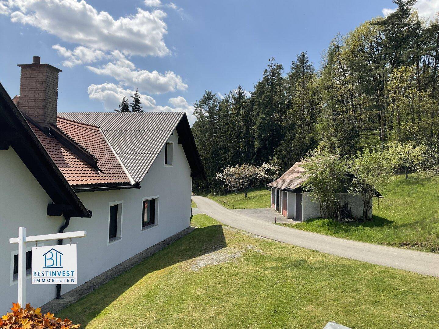 Seitenansicht des Hauses mit einem kleinen Nebengebäude und einer Zufahrt, umgeben von Bäumen.