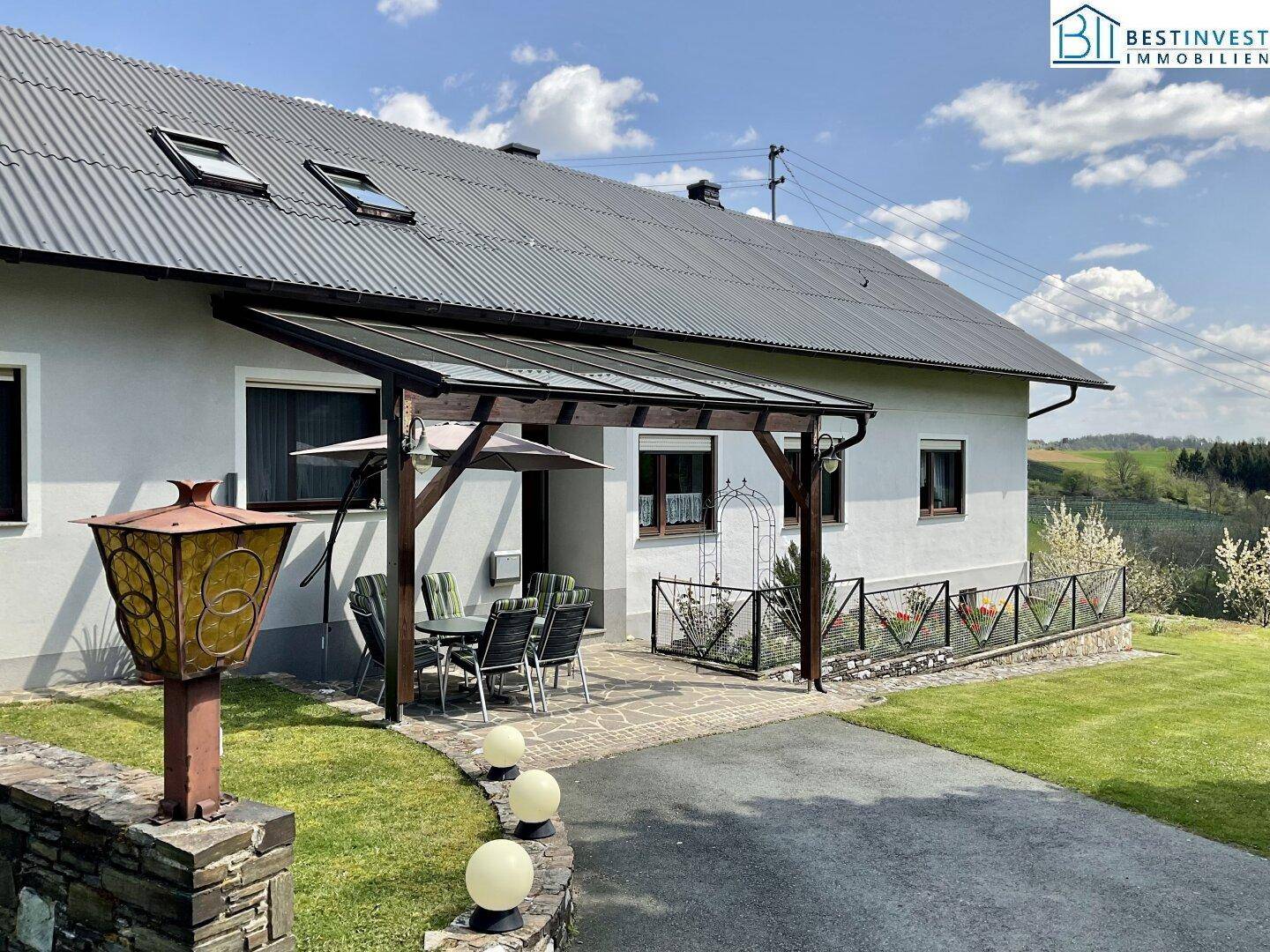 Überdachte Terrasse mit Gartenmöbeln am Haus, umgeben von grüner Landschaft unter blauem Himmel.