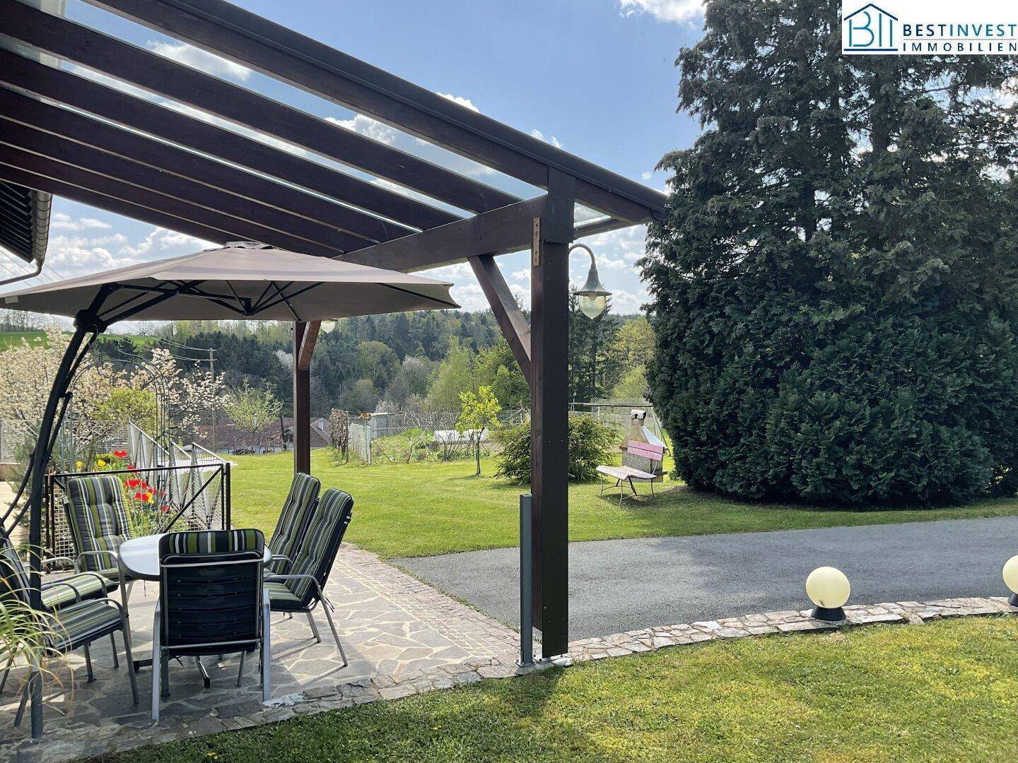 Gemütliche überdachte Terrasse mit Esstisch und Sonnenschirm, angrenzend an den grünen Gartenbereich.
