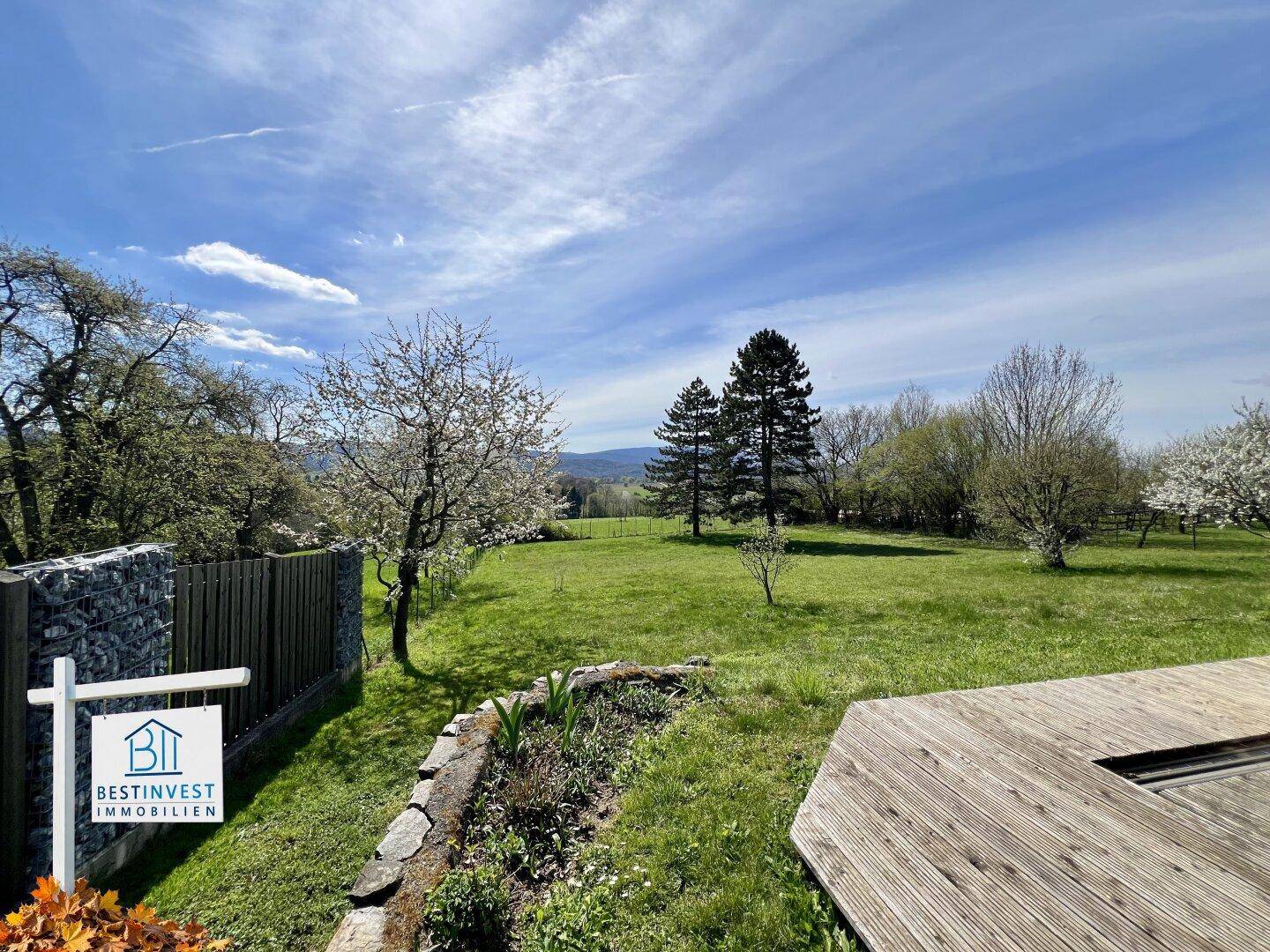 Gepflegter Garten mit Holzterrasse und Blick auf die umliegende grüne Landschaft und Bäume.