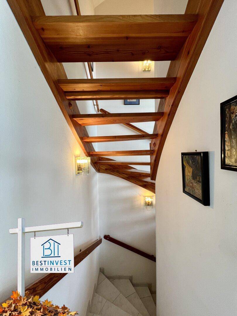 Helles Treppenhaus mit offener Holztreppe und weißen Wänden, gut beleuchtet.