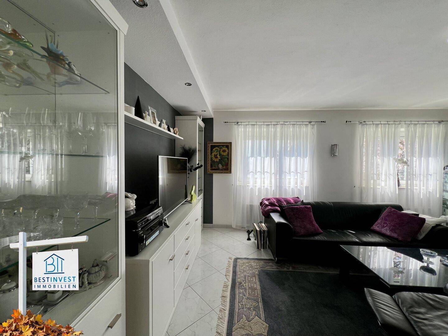 Geräumiges Wohnzimmer mit bequemer Couch, Medienschrank und hellen Fenstern, modern eingerichtet.