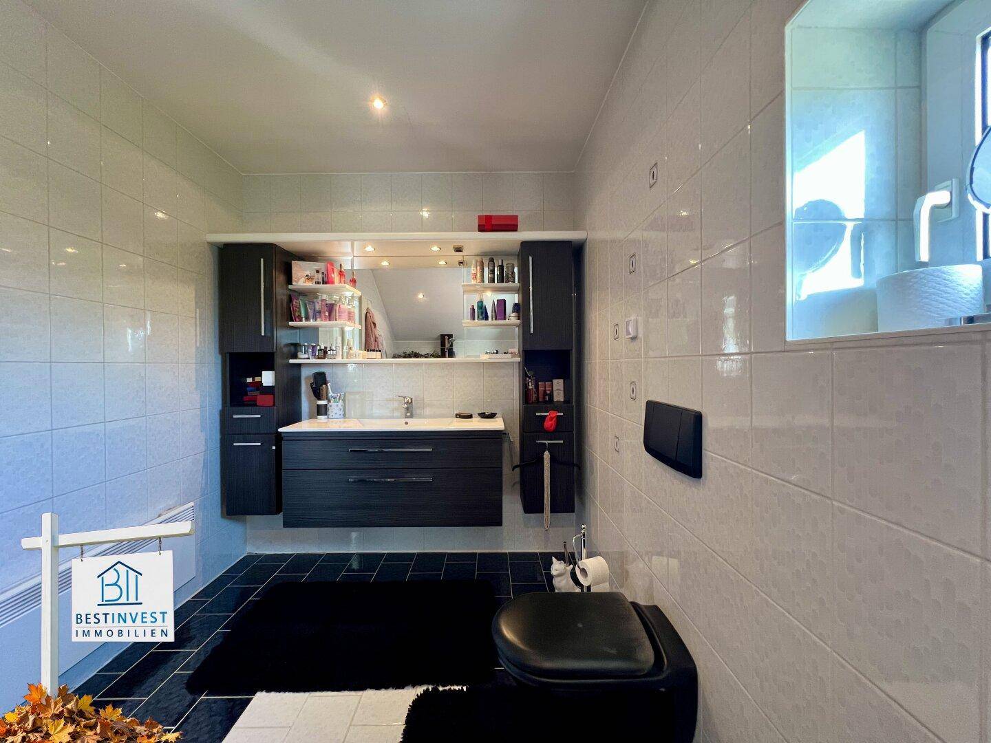 Modernes Badezimmer mit dunklem Waschtisch, WC und weißen Fliesen, gut beleuchtet durch ein Fenster.