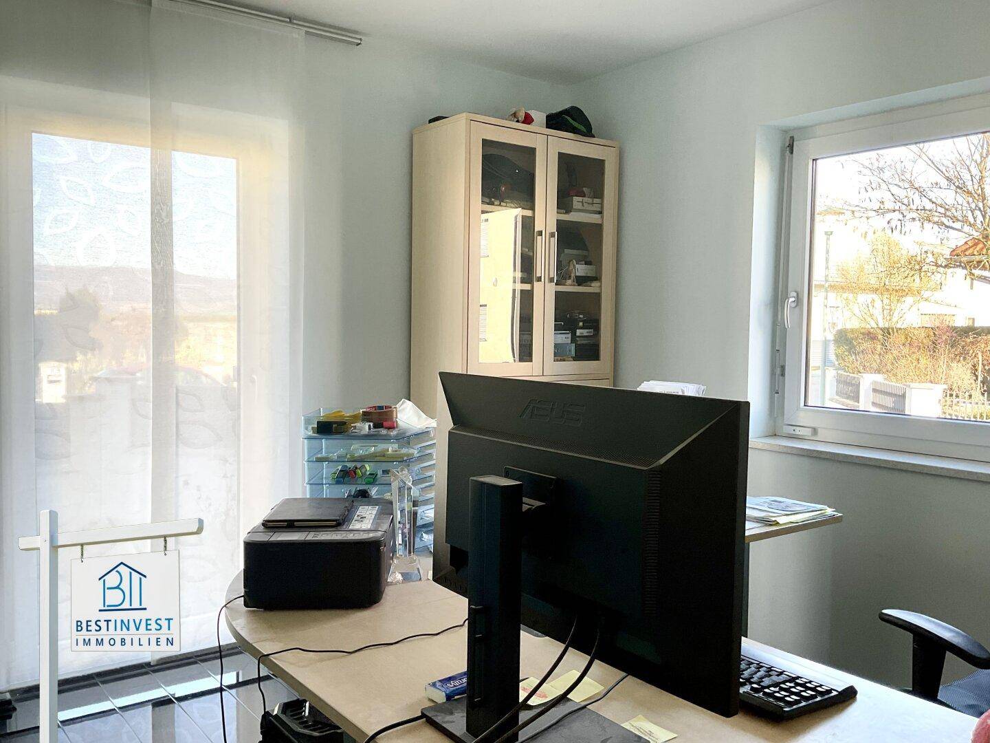 Helles Büro mit Schreibtisch, Schrank und Fenstern, die einen weiten Blick ins Grüne bieten.