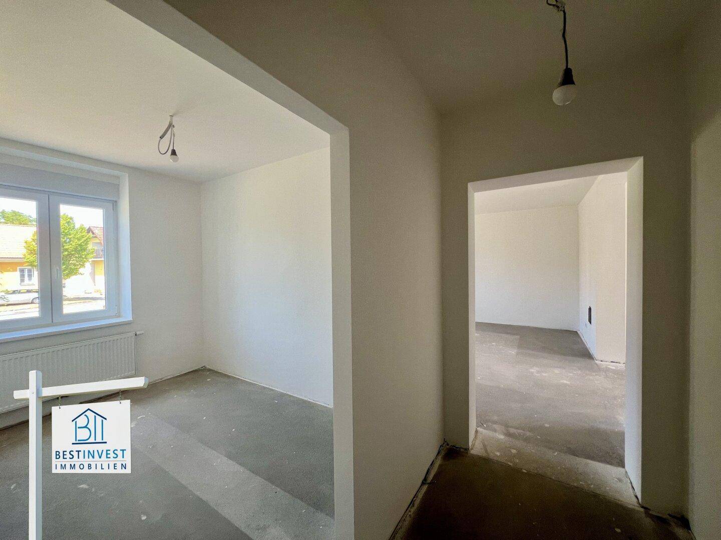 Leeres Zimmer im Rohbau mit Fenster und offenem Zugang zu einem weiteren Raum, ideal für individuelle Nutzung.