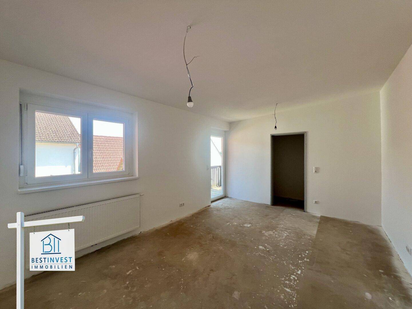Geräumiges Zimmer im Rohbau mit Fenstern und Zugang zu einem Außenbereich, ideal für Wohnzwecke.