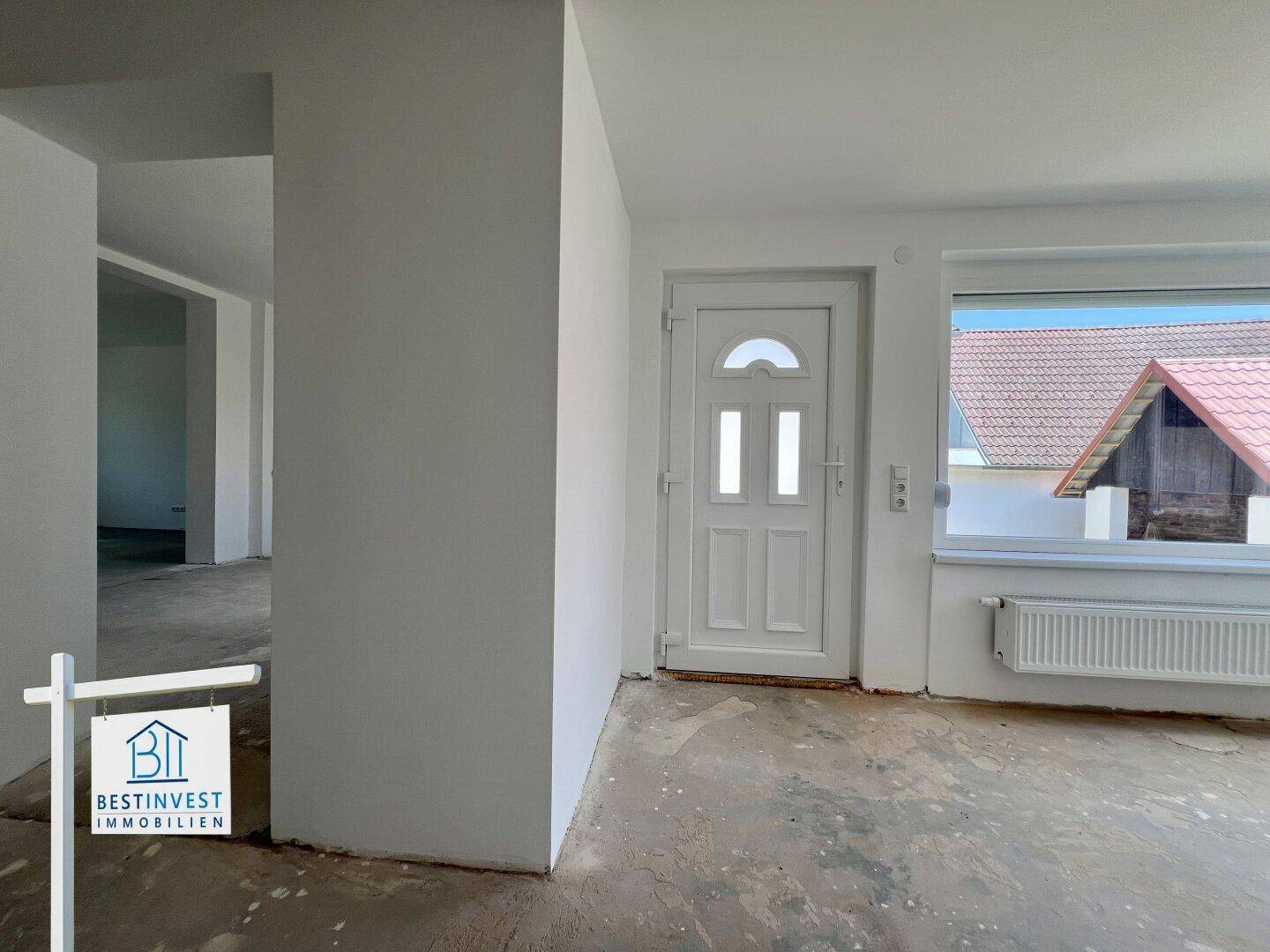 Eingangsbereich im Rohbau mit weißer Tür und großem Fenster, das viel Tageslicht hereinlässt.