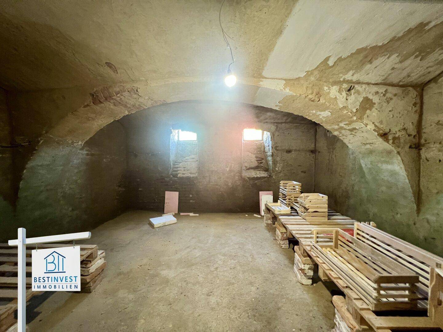 Historischer Gewölbekeller mit unverputzten Wänden, kleinen Fenstern und Holzpaletten als Lagerflächen.