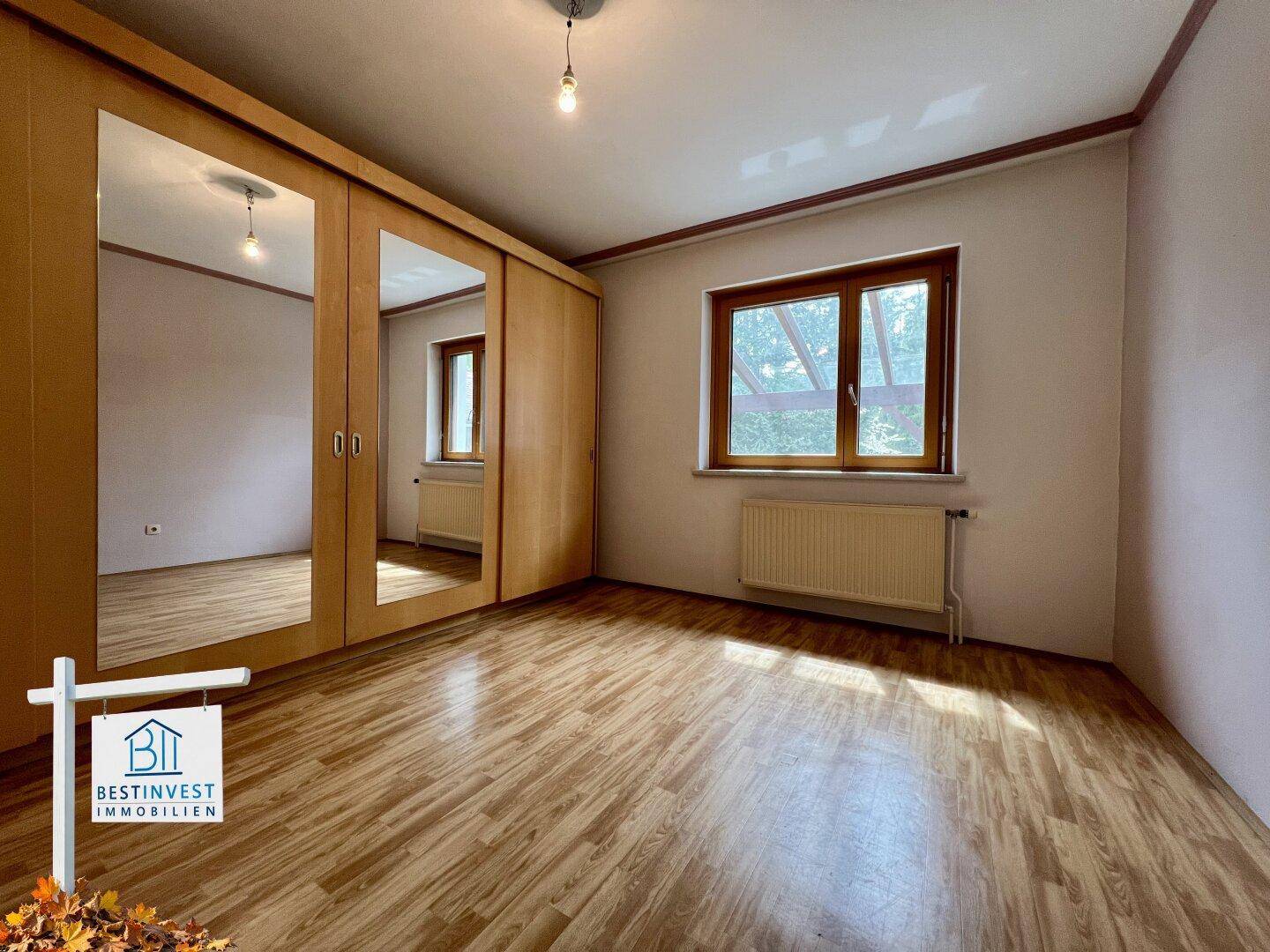 Schlafzimmer mit Laminatboden, einem großen Spiegelschrank und einem Fenster mit Blick ins Grüne.
