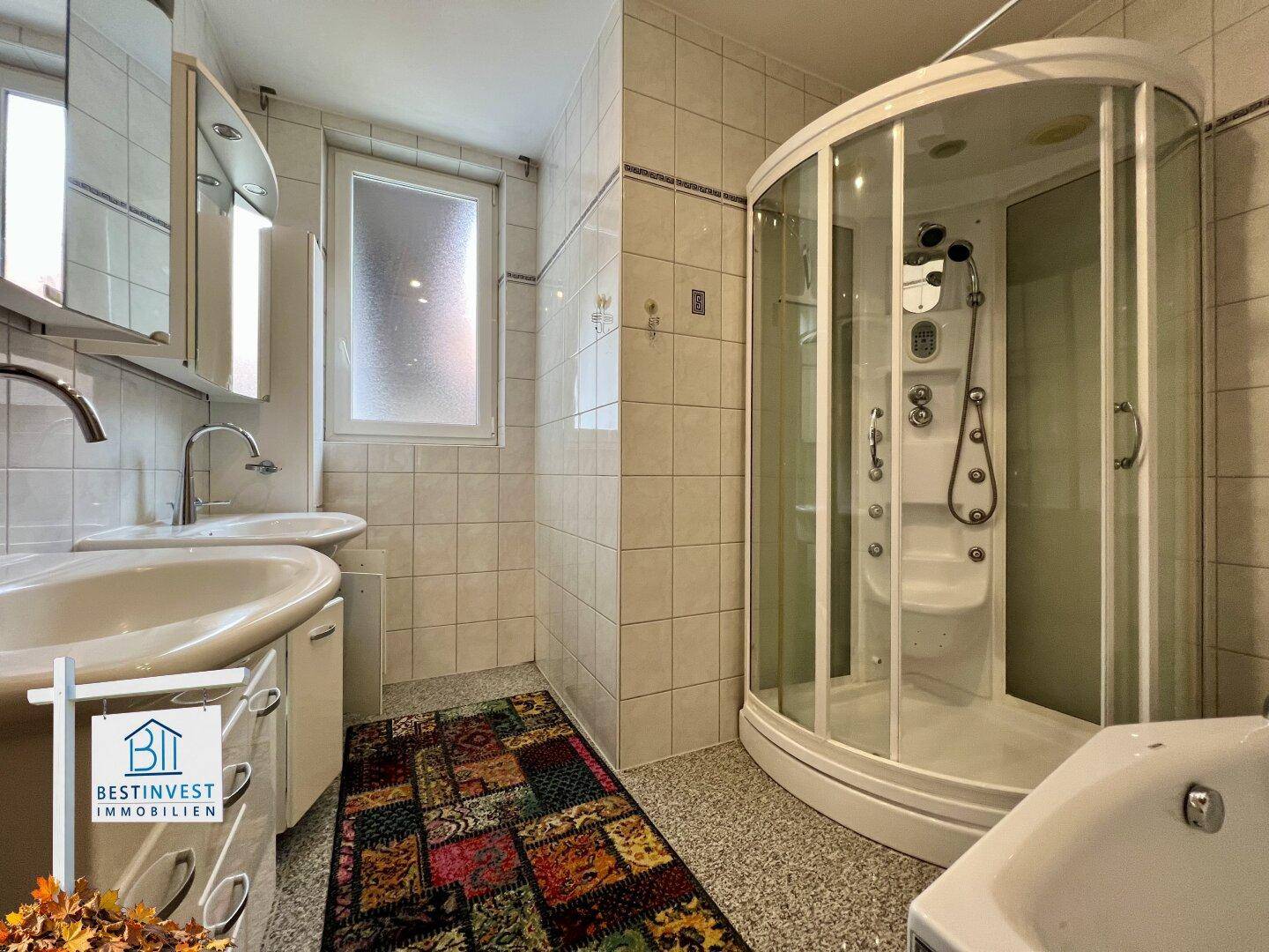 Badezimmer mit zwei Waschbecken, einer Duschkabine und hellen Fliesen an Wänden und Boden.
