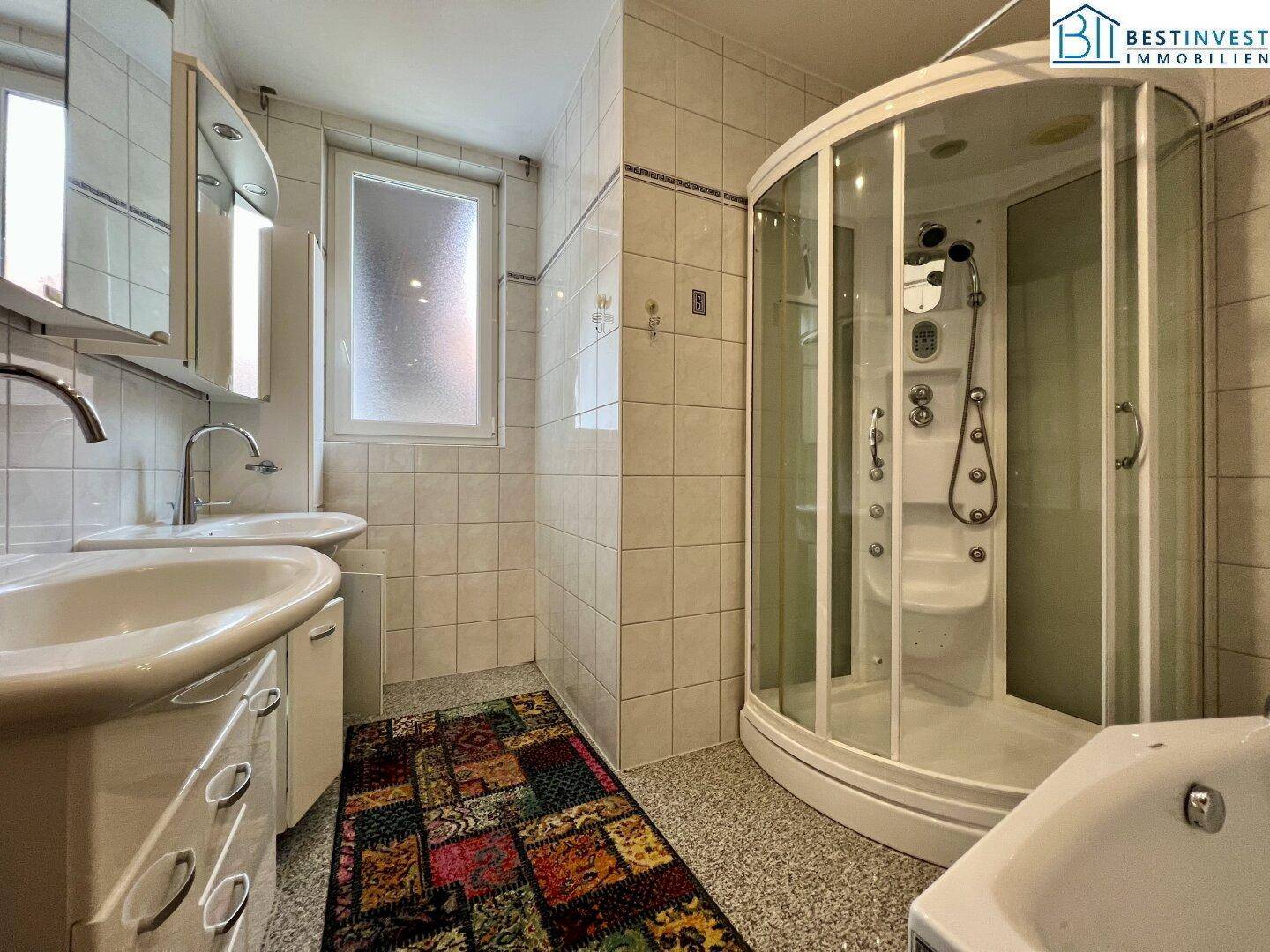 Gefliestes Badezimmer mit zwei Waschbecken, einer Duschkabine und einem Fenster für Tageslicht.