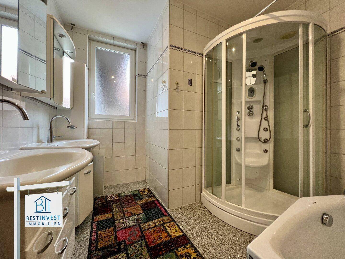 Badezimmer mit Duschkabine, zwei Waschbecken und Fenster, hell gefliest.