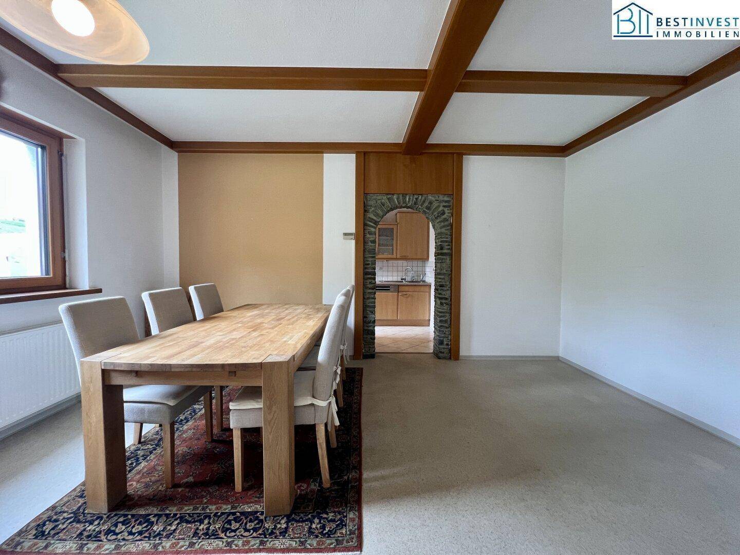 Gemütlicher Essbereich mit Holzbalkendecke und Teppichboden, verbunden mit der Küche durch einen rustikalen Steinbogen.