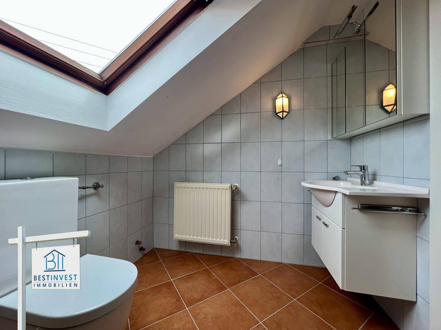 Helles Badezimmer mit WC, Waschbecken, Spiegelschrank und Heizkörper unter dem Dachfenster.