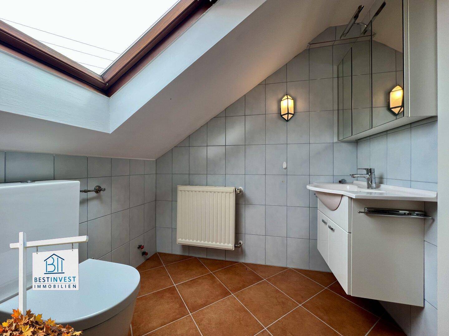 Dachgeschoss-Badezimmer mit WC, Waschbecken, Spiegelschrank und Heizkörper unter dem Dachfenster.