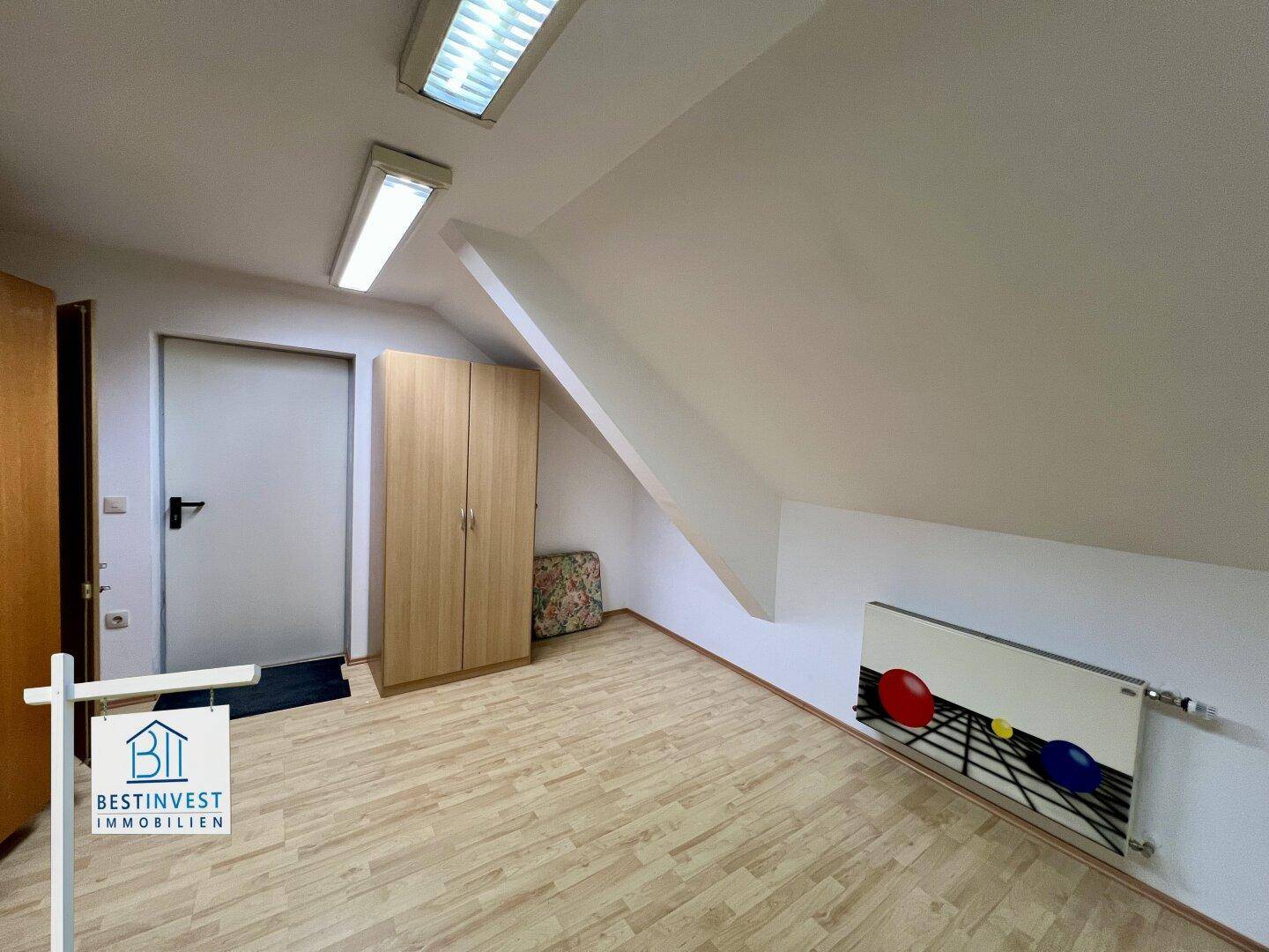 Zimmer mit Dachschrägen, Laminatboden und einem Heizkörper mit farbenfrohem Design.