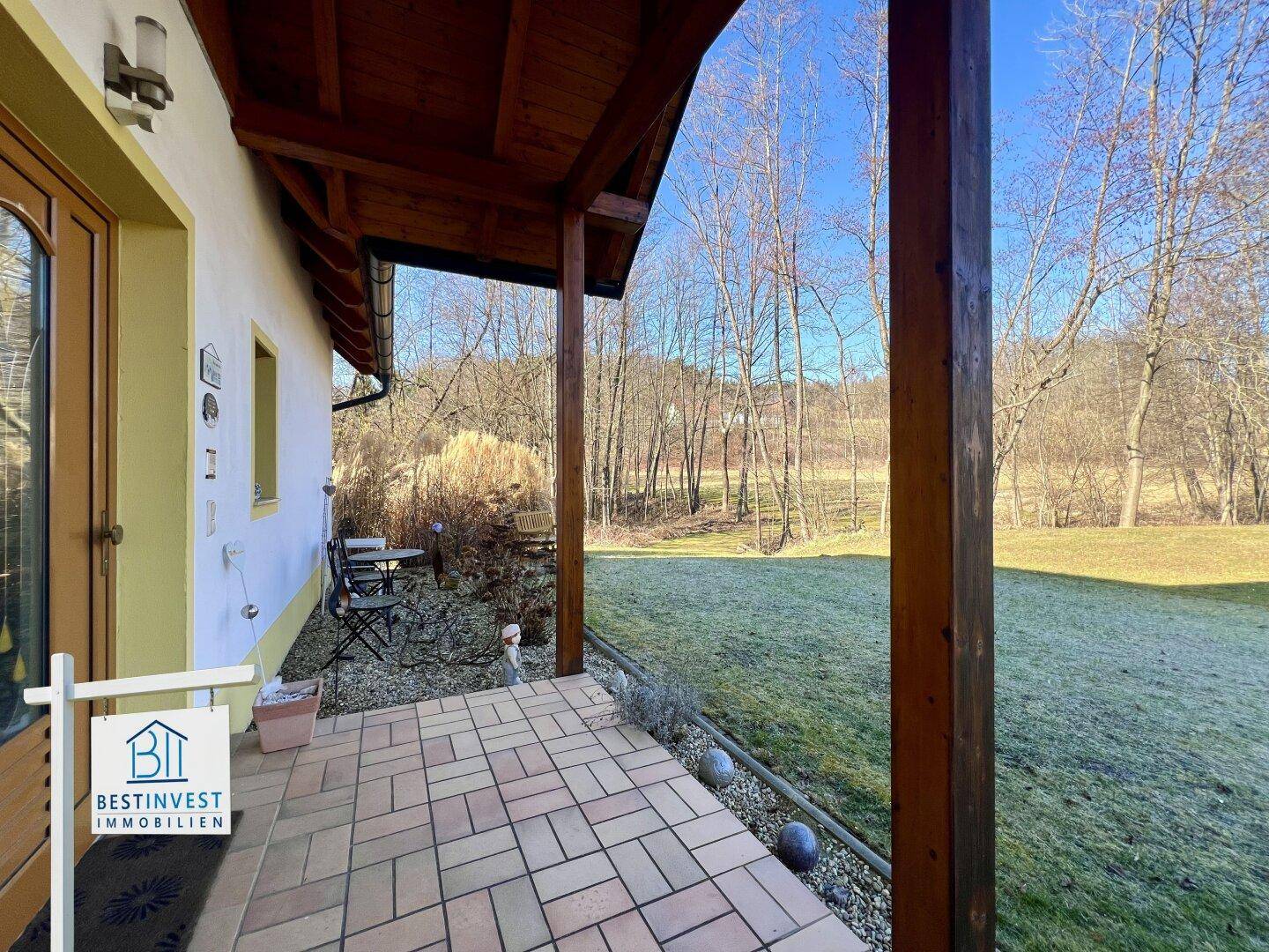 Überdachte Terrasse mit Fliesenboden und Holzbalken, die einen Blick in den Garten bietet.