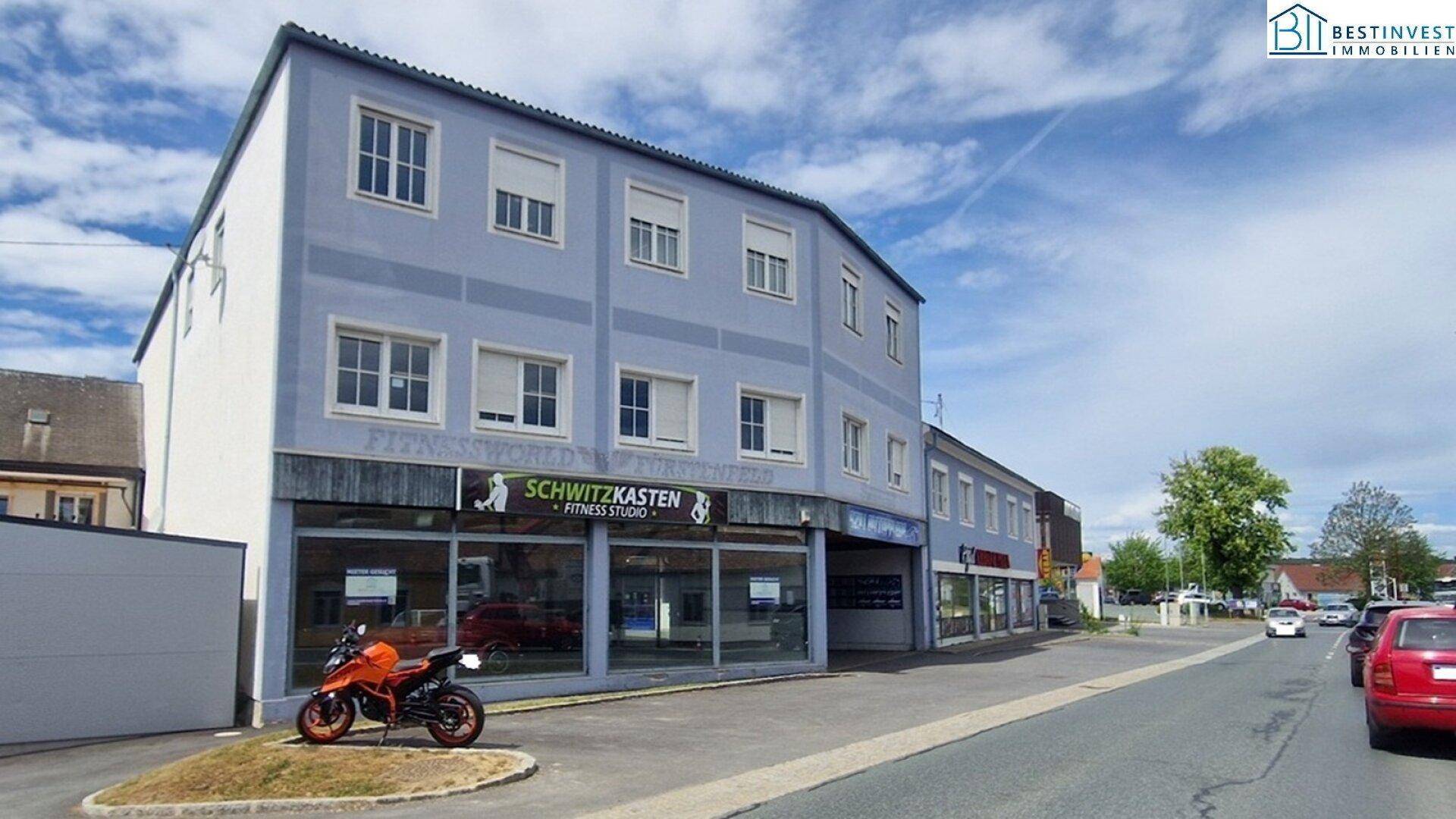 Außenansicht eines Gebäudes mit blauer Fassade und Schaufenstern, die ein Fitnessstudio zeigen, ein Motorrad parkt davor.