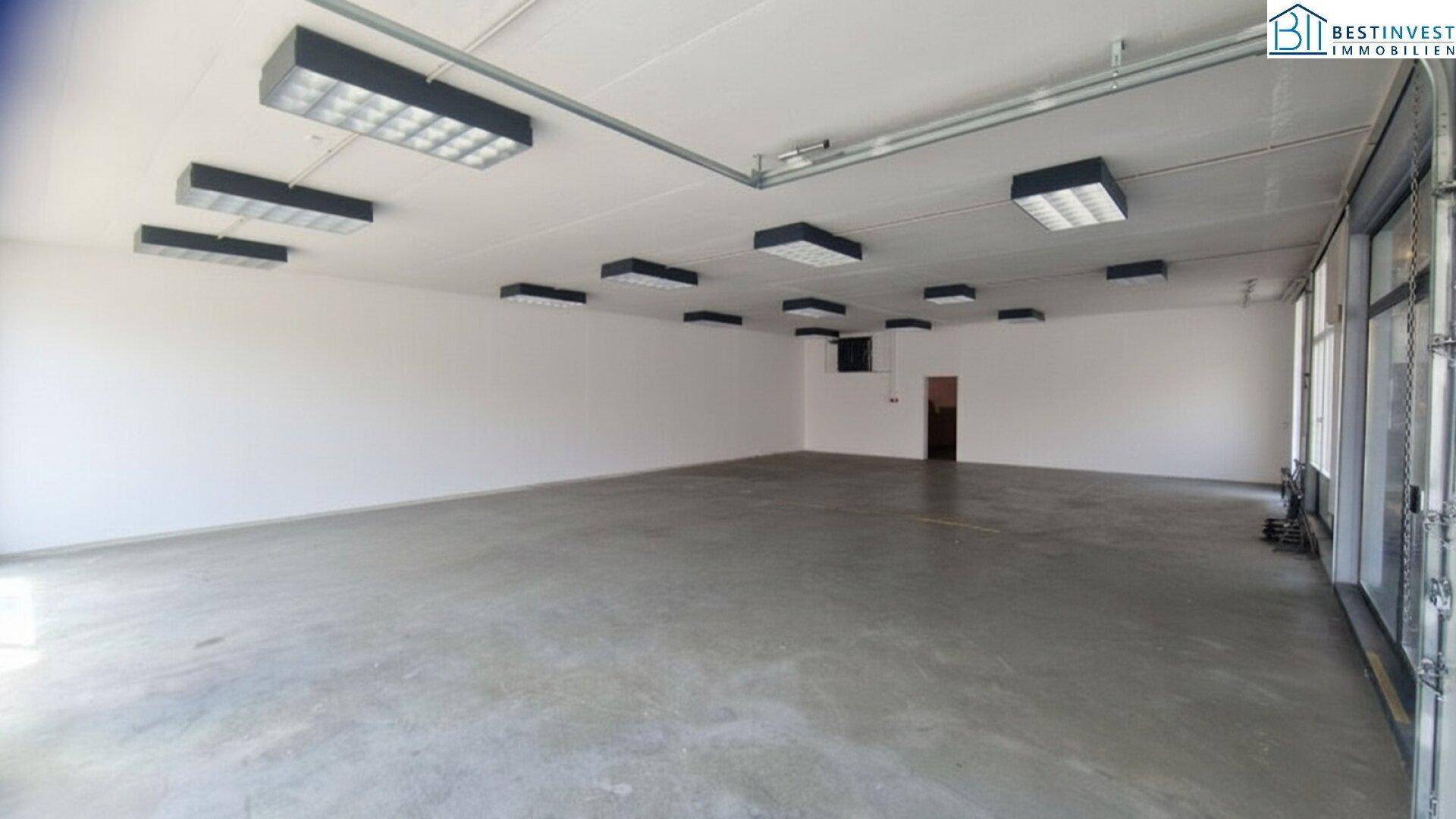 Geräumiger, leerer Gewerberaum mit Betonboden, Rolltor und Zugang zu weiteren Räumlichkeiten, bereit zur individuellen Gestaltung.
