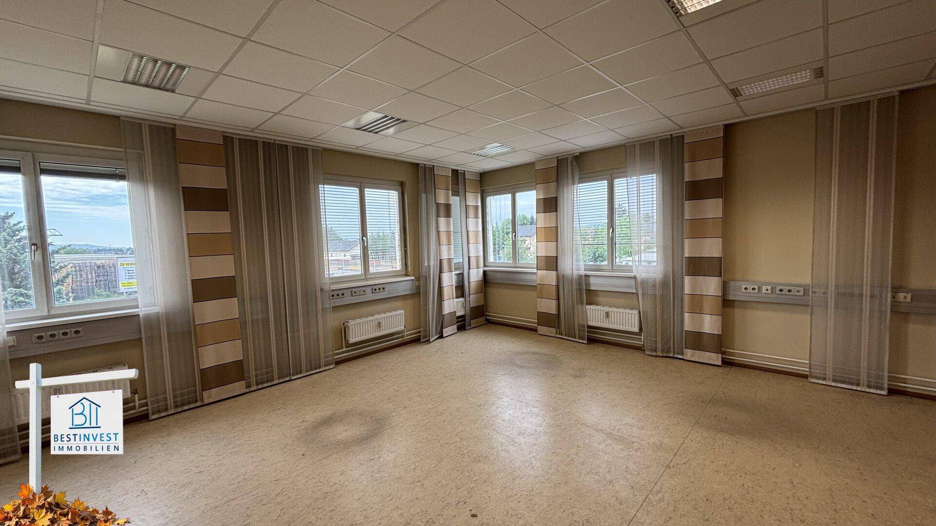 Leeres Büro mit mehreren Fenstern, Heizkörpern und gestreiften Vorhängen.