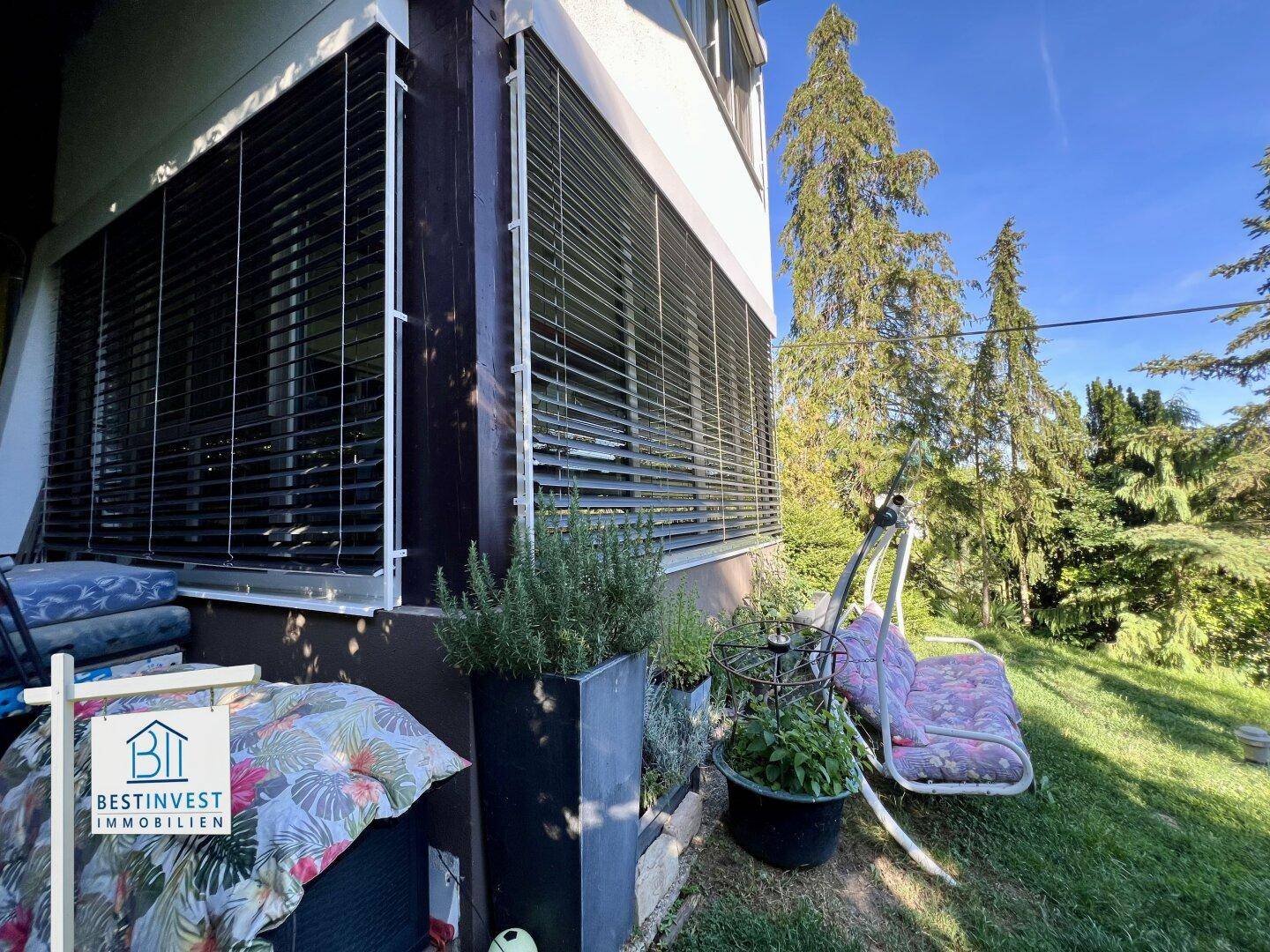 Seitlicher Bereich des Hauses mit Außenjalousien, Pflanzenkübeln und einer Gartenschaukel.