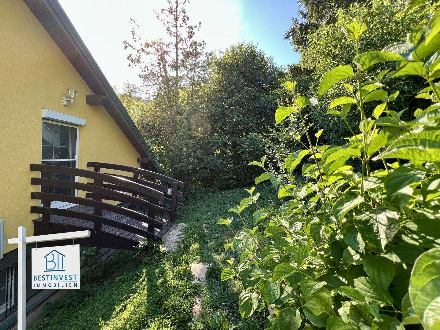 Seitlicher Hauszugang mit Holzgeländer, umgeben von üppigem Grün und Bäumen.