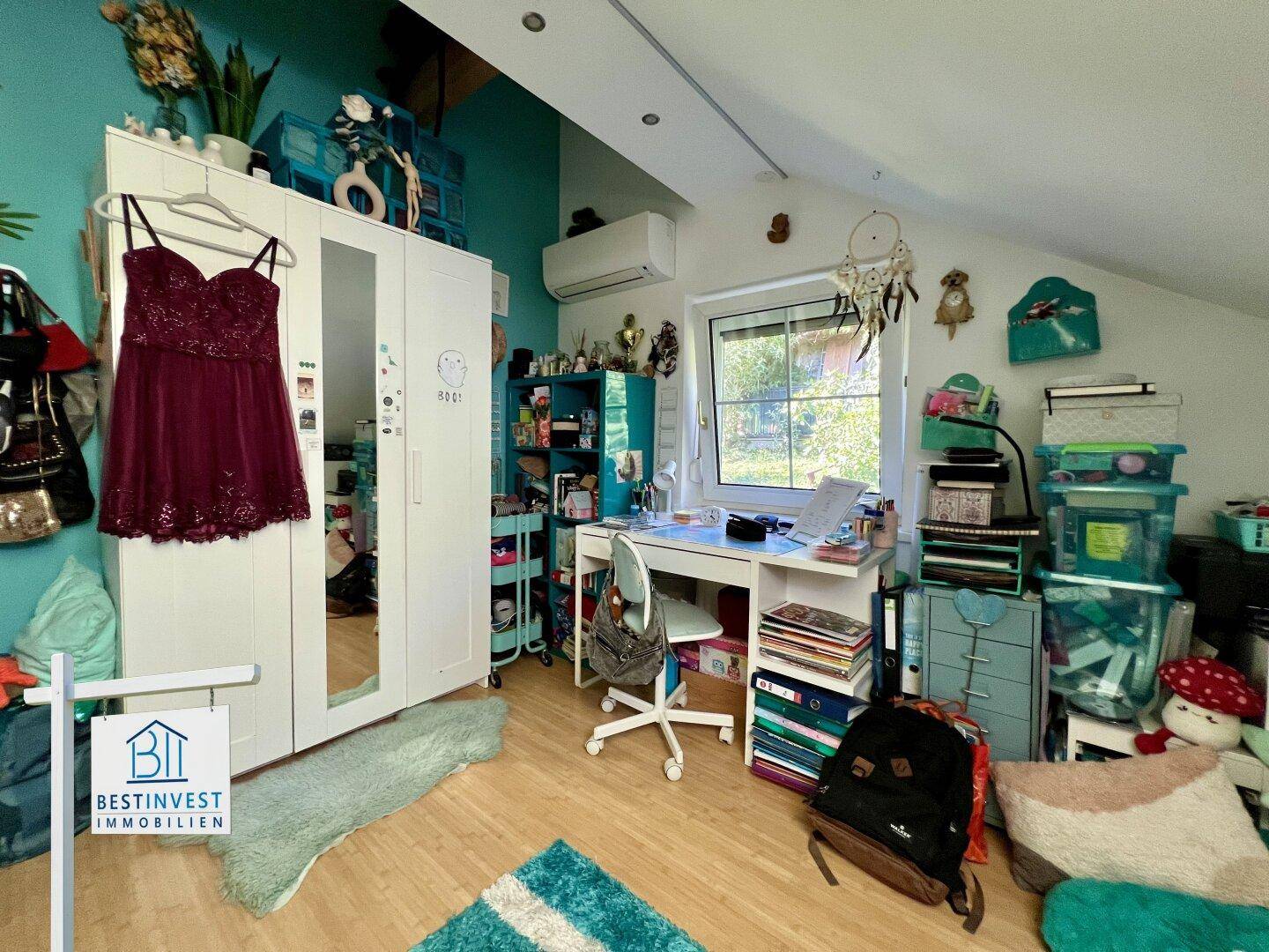 Helles Zimmer mit Schreibtisch, Kleiderschrank und schrägen Wänden unter dem Dach.