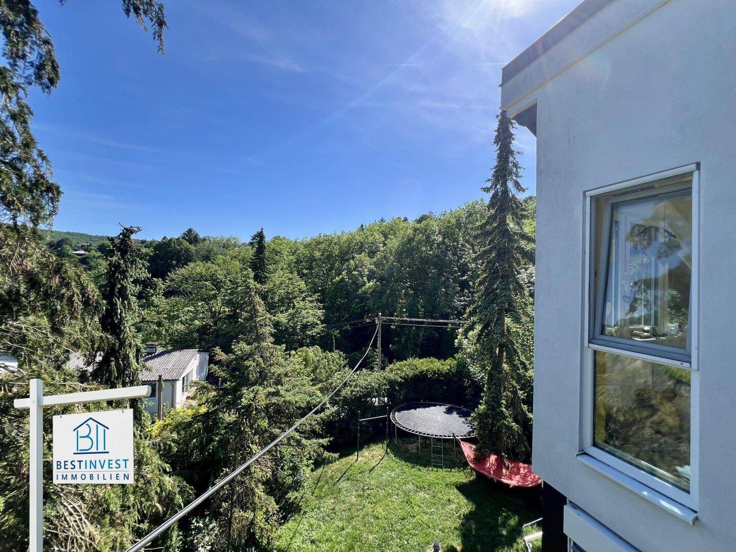 Gartenbereich mit Trampolin, umgeben von hohen Bäumen und einem sonnigen Himmel.