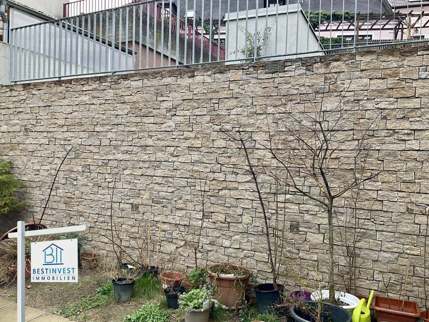 Steinmauer im Außenbereich mit verschiedenen Topfpflanzen und jungem Bewuchs.