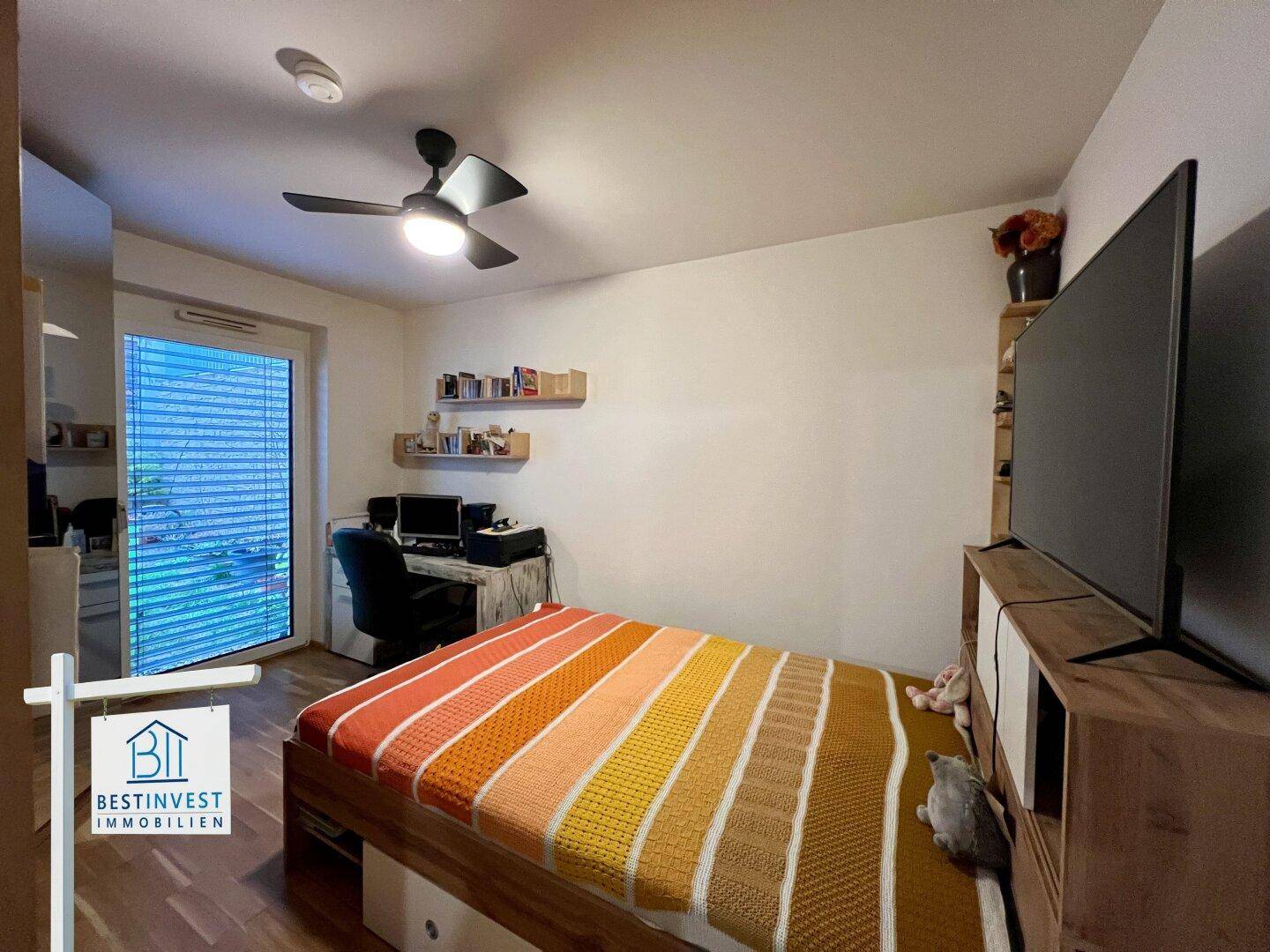 Helles Schlafzimmer mit Holzboden, Deckenventilator und Schreibtischbereich.
