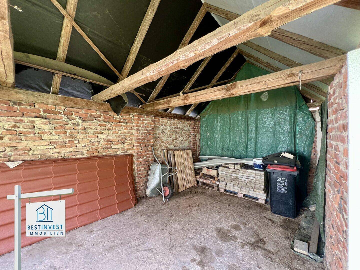 Rustikaler Innenraum eines Nebengebäudes mit unverputzten Ziegelwänden, Holzbalken und gelagerten Materialien.