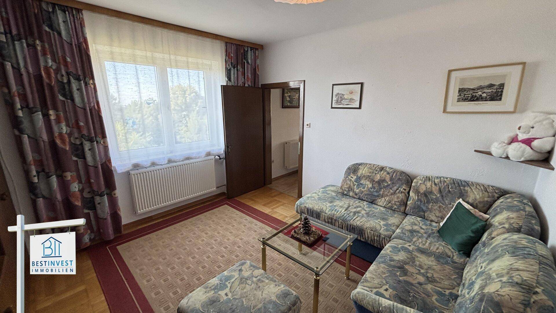 Gemütliches Wohnzimmer mit Ecksofa, Teppichboden und Blick auf die grüne Umgebung.