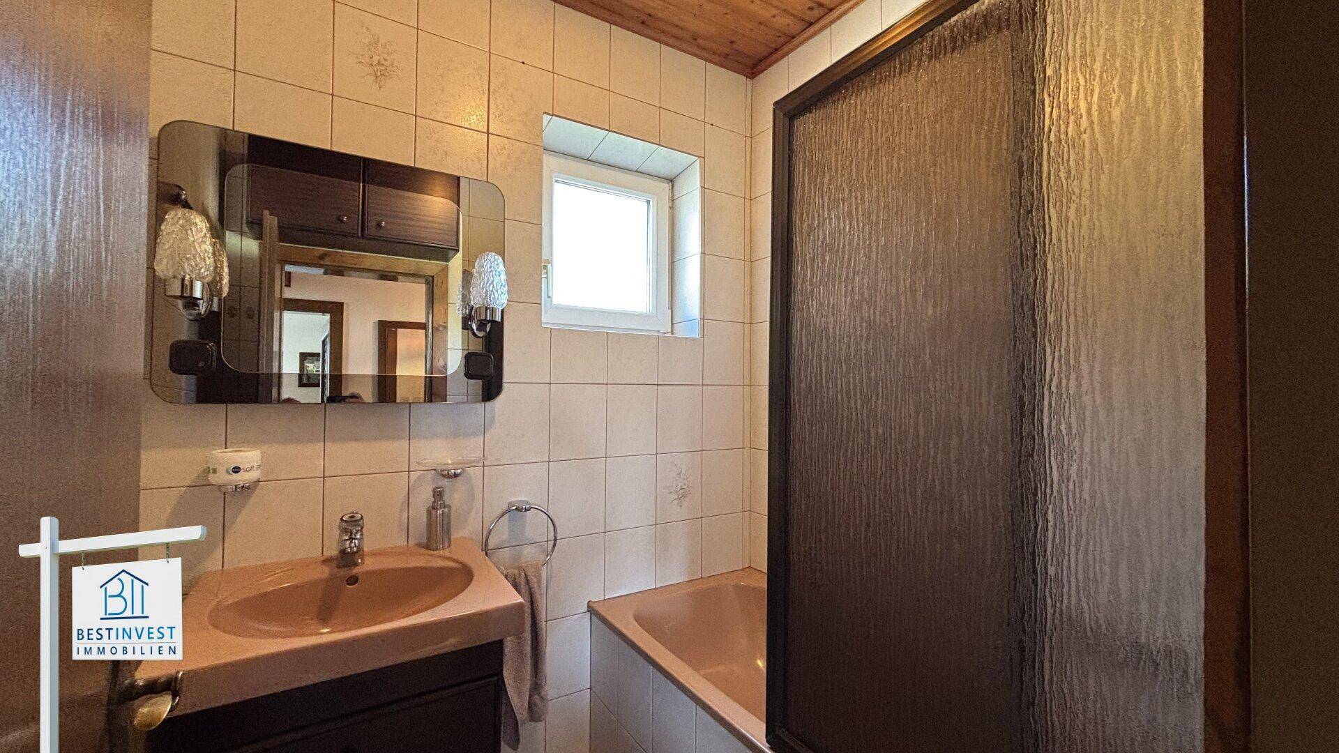 Badezimmer mit beigen Fliesen, Waschbecken und Duschkabine, mit Fenster.